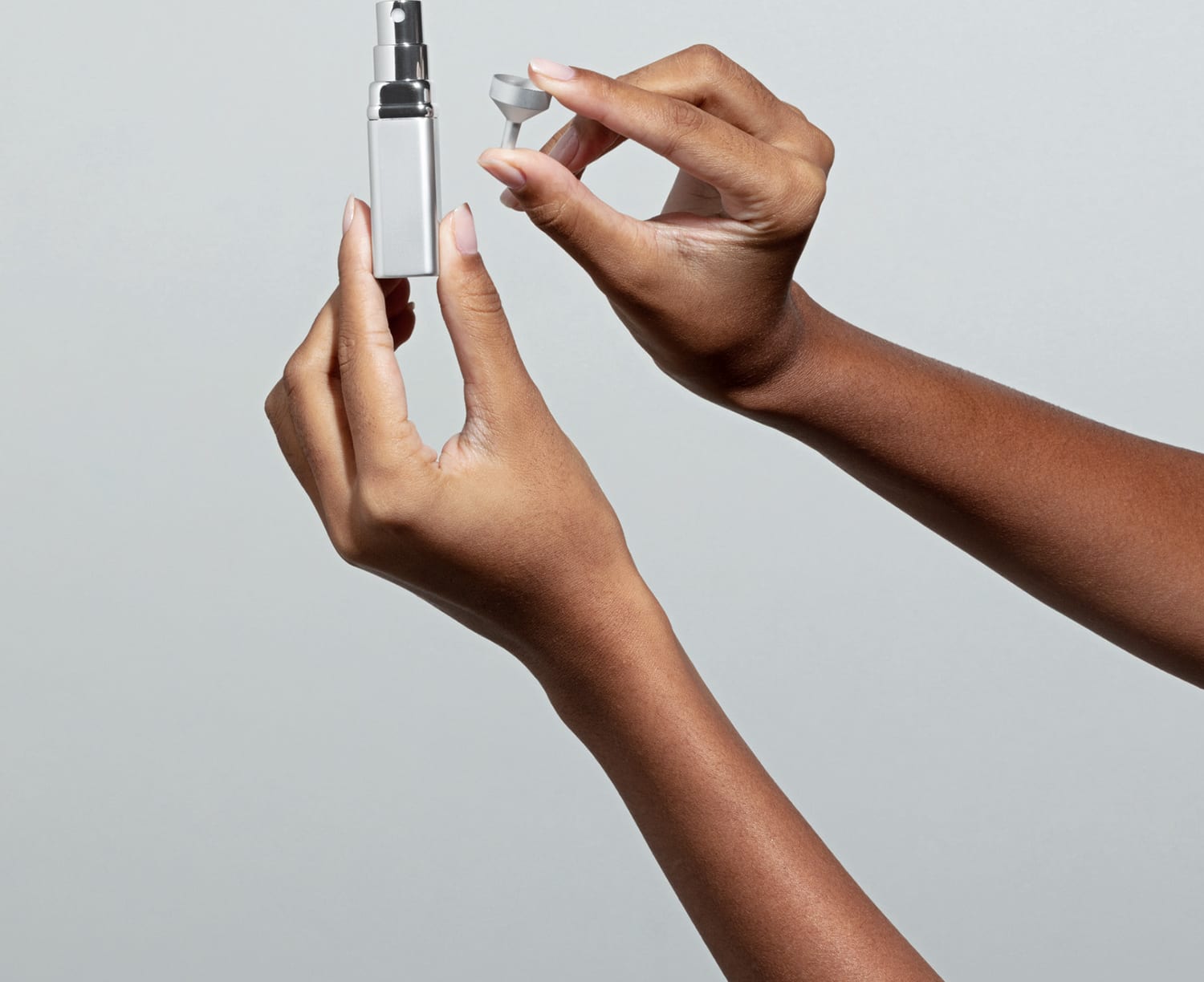 Uma mão feminina está segurando um porta-perfume com vaporizador em tubo prateado e a outra mao segura um mini funil em fundo azul claro.