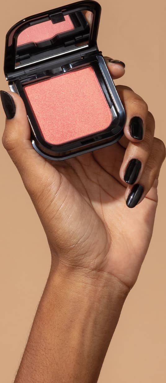 Uma mão feminina com unhas pintadas de preto está segurando um blush em pó mate Luminoso Rose Make B. na cor rosa em estojo quadrado preto de cantos arredondados e espelho na tampa. O fundo da imagem é em tons de marrom.