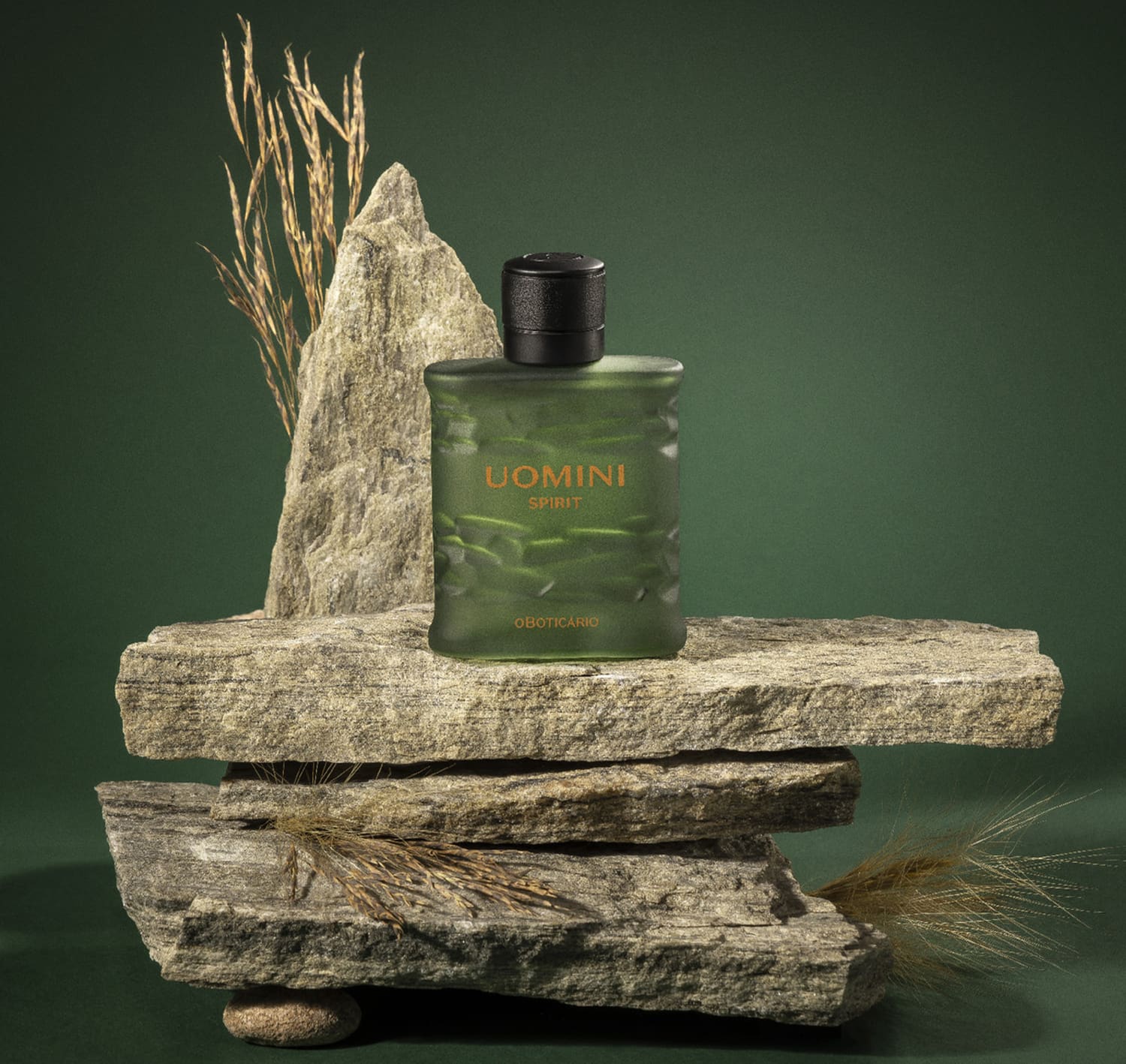 Frasco retangular em tom de verde oliva com tampa texturizada preta e letras laranjas contendo 100ml de desodorante colônia Uomini Spirit sobre superfície rochosa com galhos de trigo, em ambiente com tons de verde escuro.