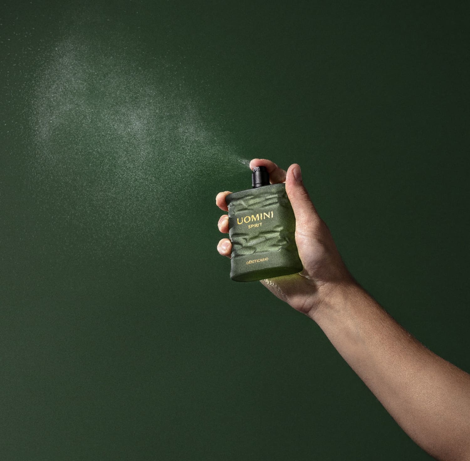 Mão segurando um frasco retangular em tom de verde oliva com tampa texturizada preta e letras laranjas contendo 100ml de desodorante colônia Uomini Spirit, espirrando o produto no ar em fundo verde bem escuro.