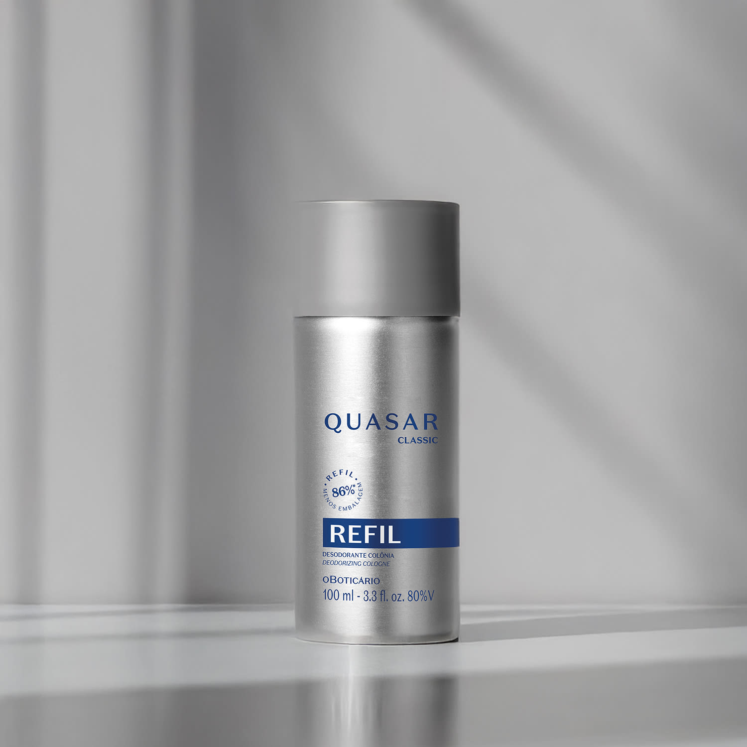 Refil de desodorante colônia Quasar Classic em frasco prateado com detalhes e letras azuis em fundo em tons de cinza com luzes naturais.