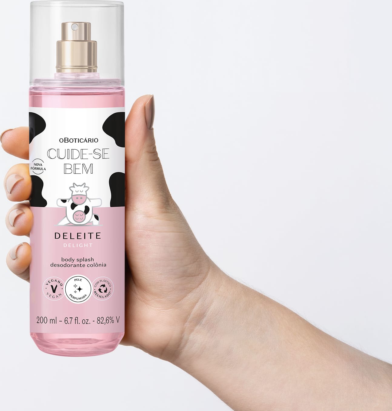 Mão feminina segurando um frasco transparente com líquido rosa, pulverizador prateado, tampa transparente e rótulo branco e rosa com manchas pretas e ilustração de vaca contendo body splash Cuide-se Bem Deleite em fundo azul.
