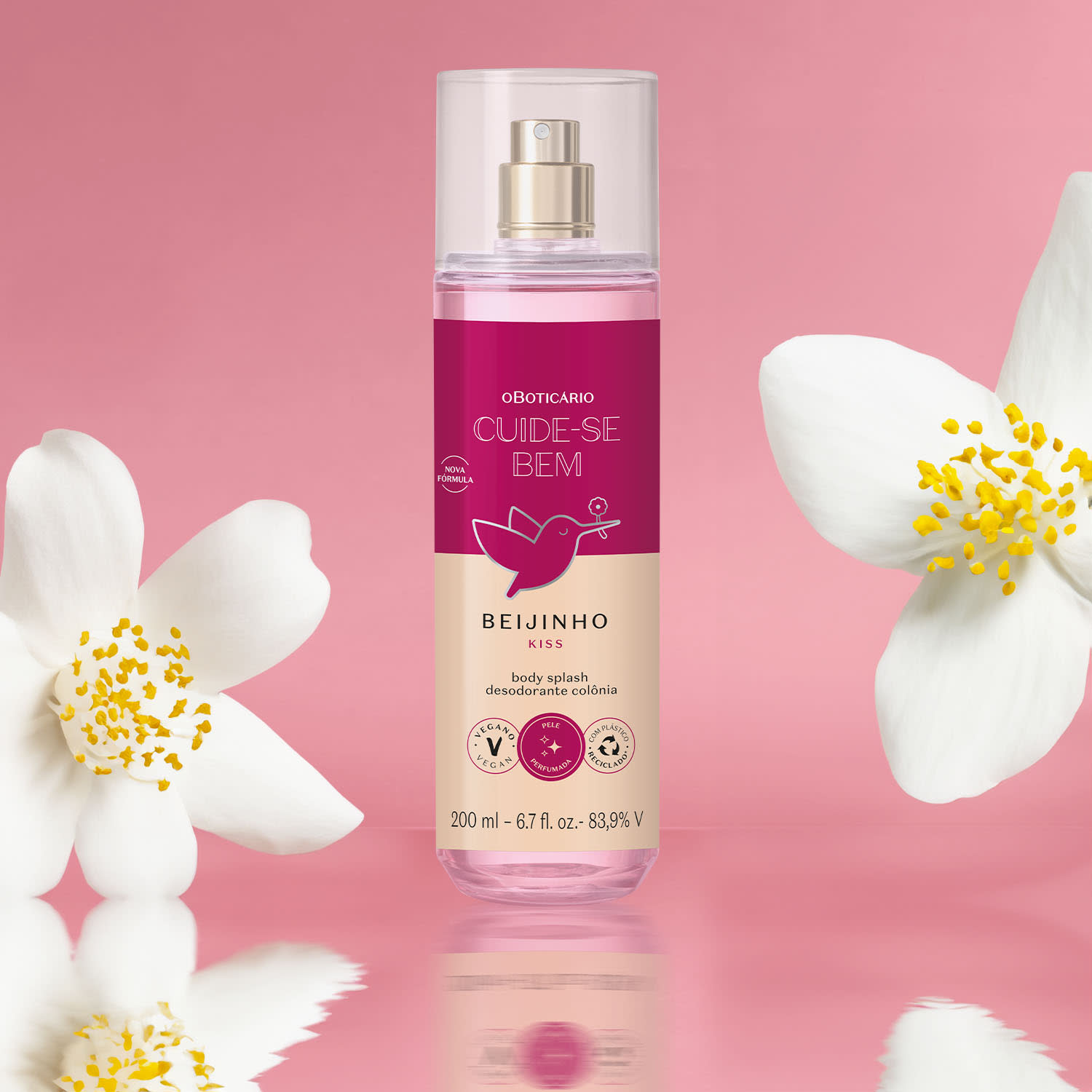 Frasco com líquido rosa, pulverizador prateado, tampa transparente e rótulo vinho e rosa com beija-flor ilustrado de body splash da marca Cuide-se Bem Beijinho em fundo rosa, com flores brancas ao redor, refletido em superfície brilhante.