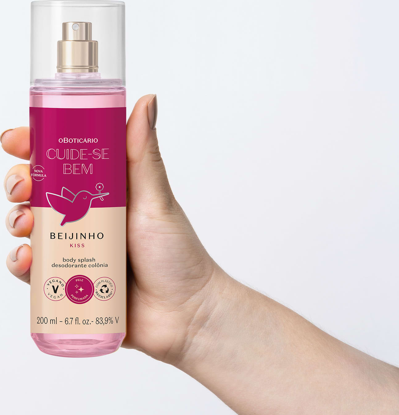 Mão segurando um frasco com líquido rosa, pulverizador prateado, tampa transparente e rótulo vinho e rosa com beija-flor ilustrado de body splash da marca Cuide-se Bem Beijinho com fundo azul claro.
