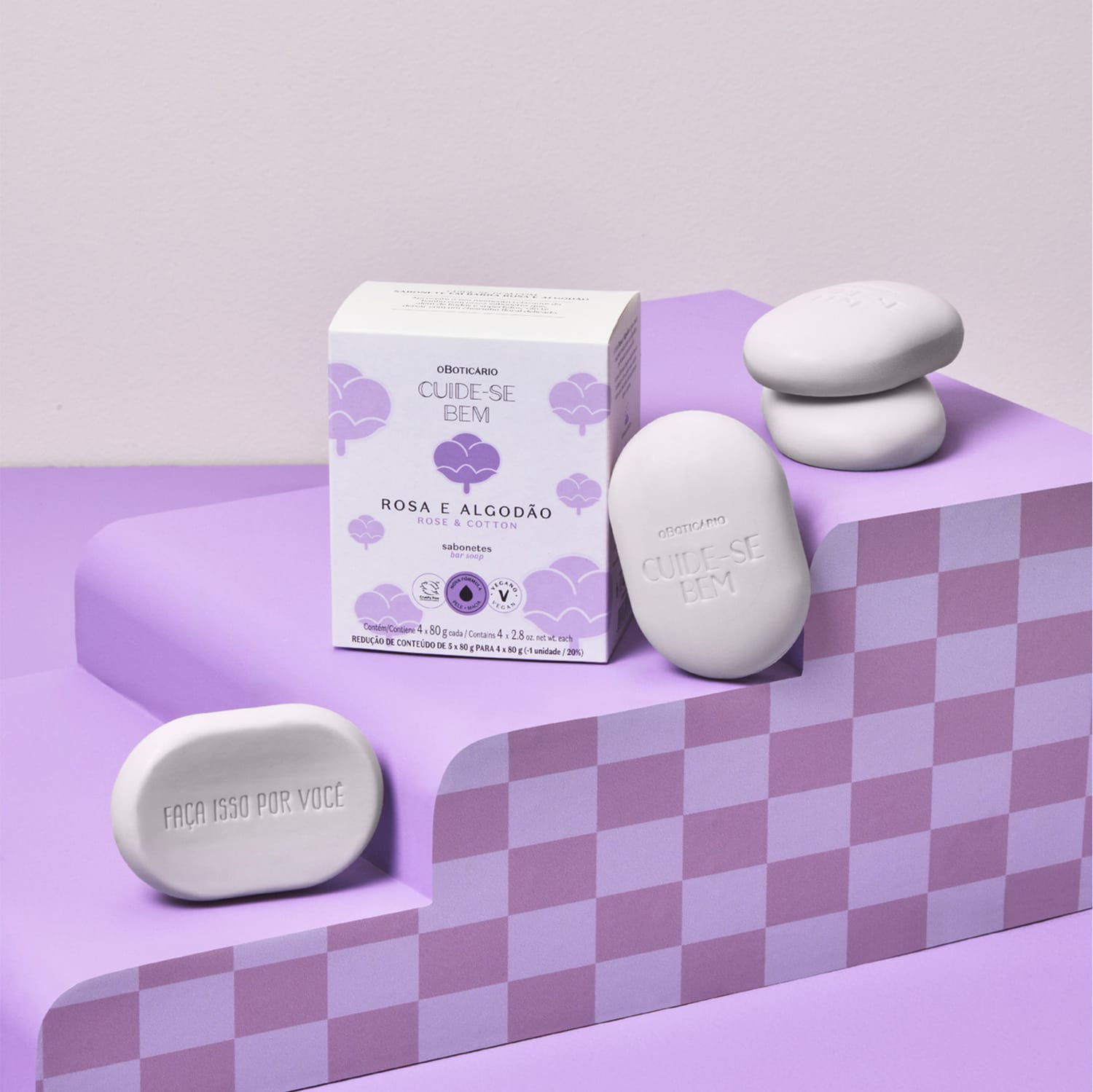 Quatro sabonetes em barra na cor lilás da marca Cuide-se Bem Rosa e Algodão ao lado de sua caixa branca e lilás em superfície em tons de roxo e fundo lilás.