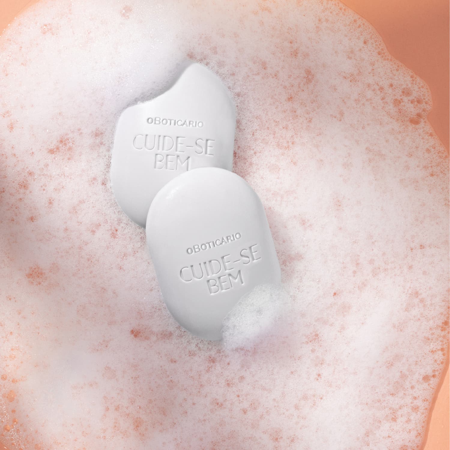 Dois sabonetes em barra ovalados brancos da marca Cuide-se Bem Nuvem em meio a muita espuma branca em fundo laranja.