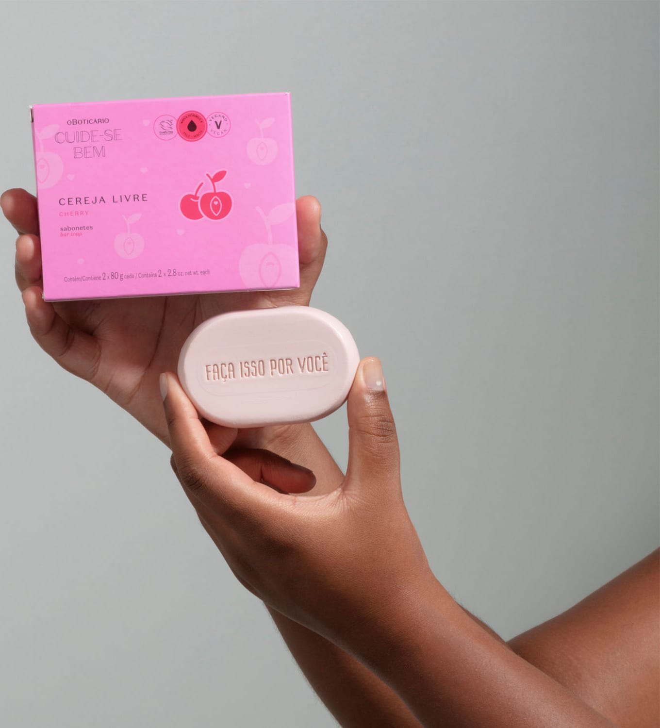 Uma mão feminina segurando uma caixa roxa com letras pretas e detalhes rosa e a outra segurando um sabonete em barra branco ovalado da marca Cuide-se Bem Cereja Livre em fundo cinza.