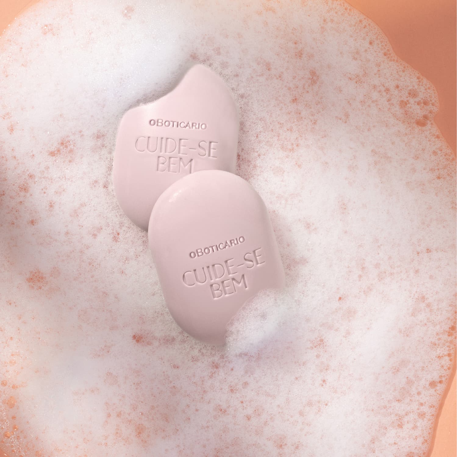 Dois sabonetes em barra brancos em formato oval da marca Cuide-se Bem Cereja Livre com muita espuma branca ao redor em fundo laranja.
