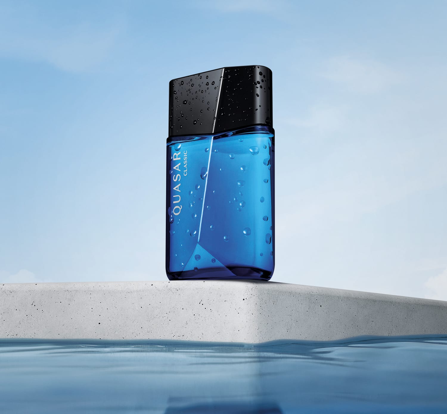Vidro azul com tampa preta, ambos em formato geométrico e letras brancas no rótulo contendo 100ml de desodorante colônia Quasar Classic flutuando sobre superfície de concreto com água ao redor, sob céu azul claro.