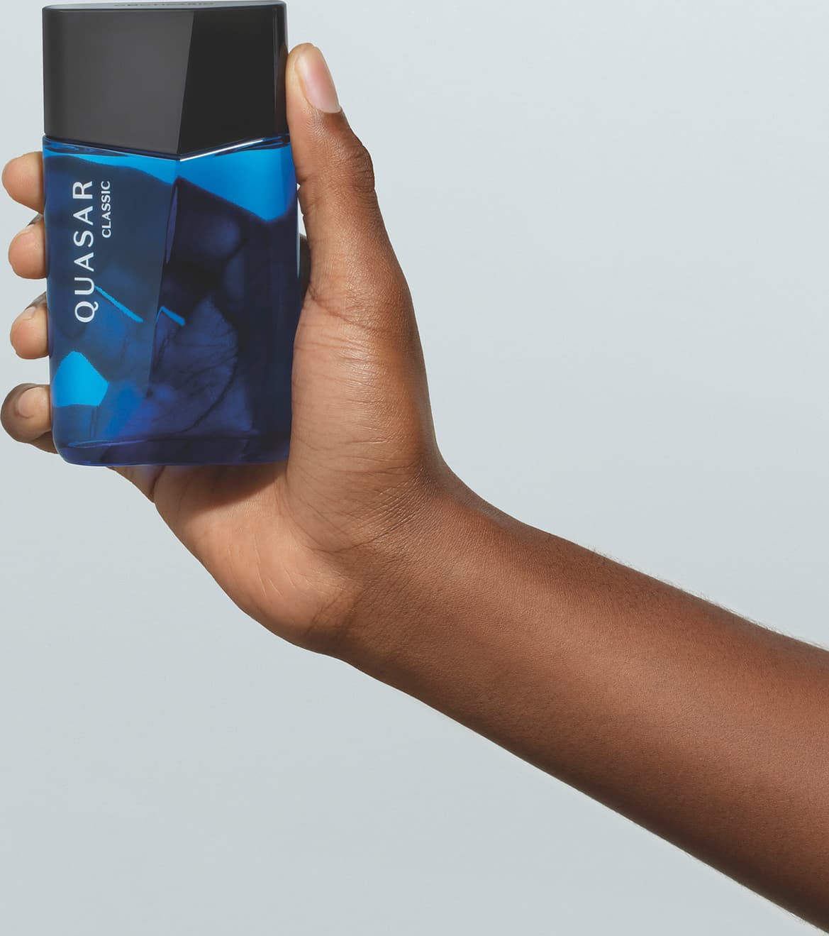 Mão masculina de pele negra segurando um vidro azul com tampa preta, ambos em formato geométrico e letras brancas no rótulo contendo 100ml de desodorante colônia Quasar Classic em fundo azul.