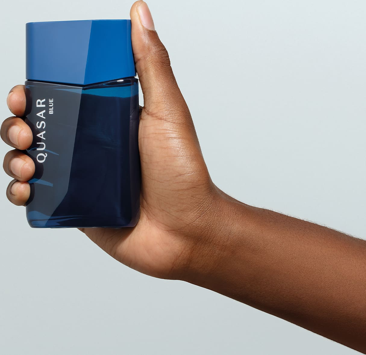 Mão masculina segurando um frasco de vidro em formato geométrico com letras amarelas contendo desodorante colônia Quasar Blue em fundo azul claro.
