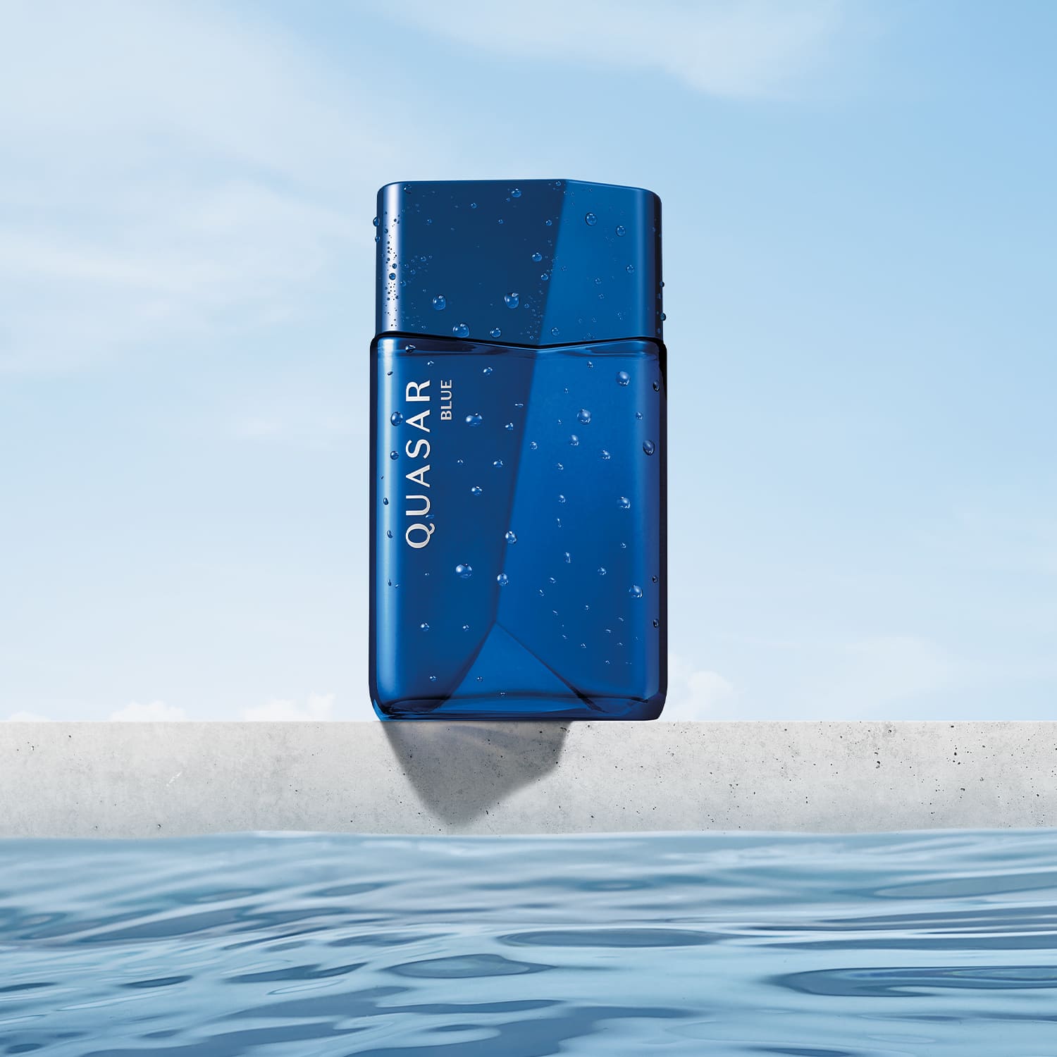 Frasco de vidro em formato geométrico com letras amarelas contendo desodorante colônia Quasar Blue com gotas d'água, flutuando sobre superfície de água em ambiente externo com céu claro.