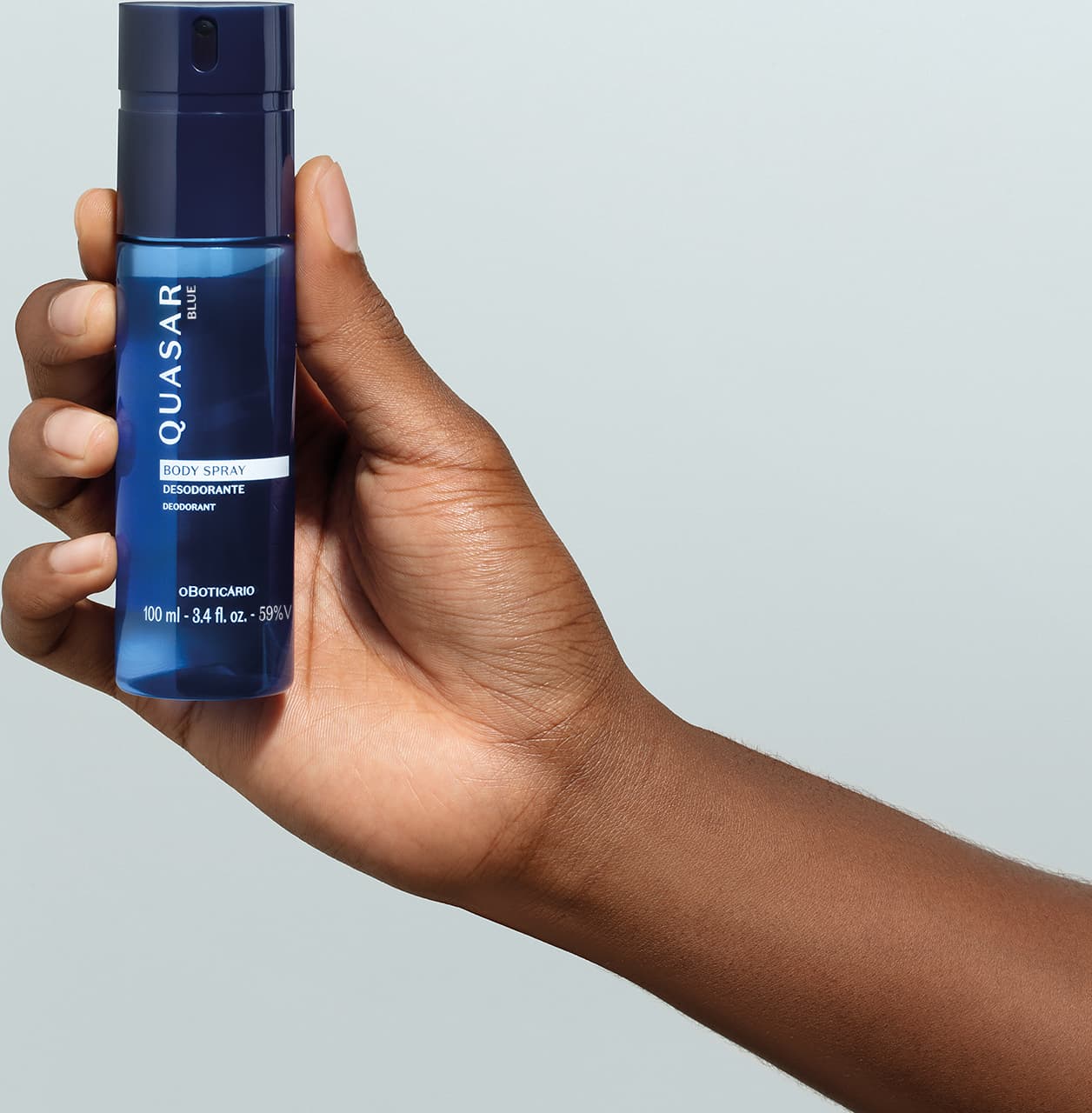 Mão masculina segurando um frasco azul marinho com letras amarelas e detalhe branco contendo body spray desodorante da marca Quasar Blue em fundo azul claro.