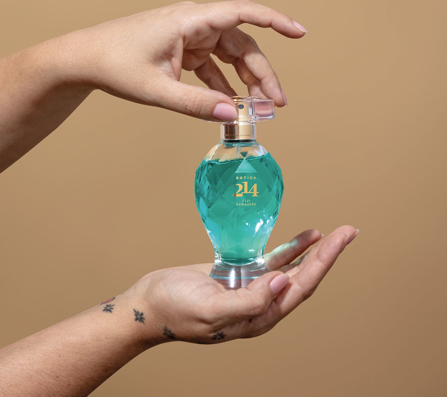 Duas mãos femininas segurando um vidro transparente lapidado com líquido azul, borrifador dourado, tampa transparente e letras douradas de Eau de Parfum Floral Especiado da marca Botica 214 Fiji Paradise em fundo marrom.