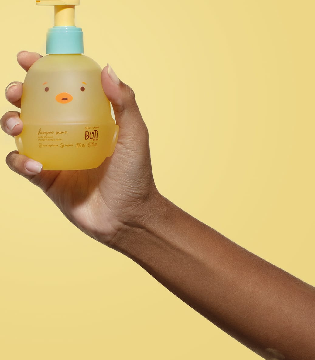 Mão segurando um frascoem forma de passarinho amarelo com detalhes laranjas, tampa azul e bomba dosadora amarela de shampoo suave da marca Boti Baby em um fundo alaranjado.