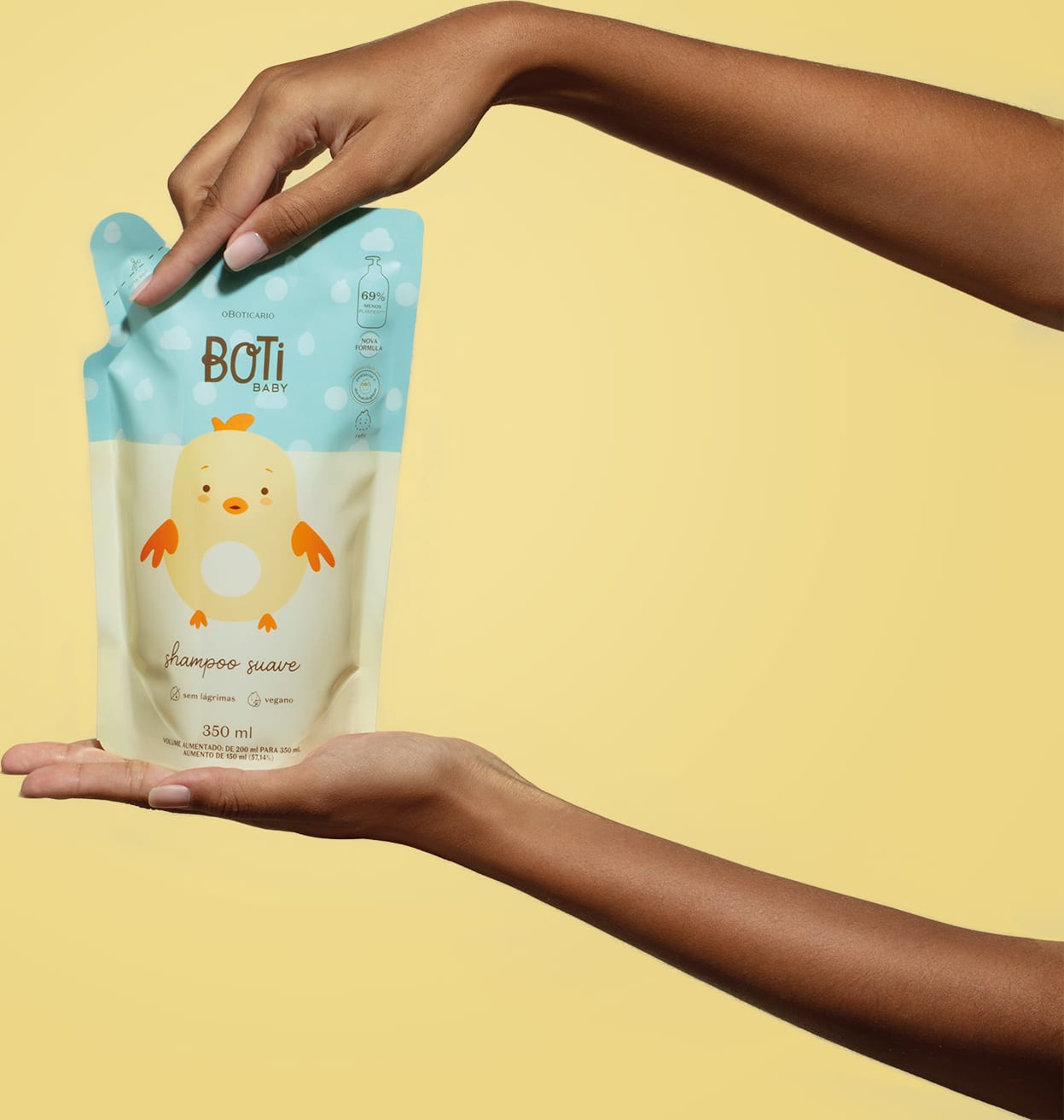 Mão segurando um Refil de shampoo suave da marca Boti Baby em sachê nas cores azul e amarelo claro com ilustração de passarinho amarelo. em um fundo alaranjado.