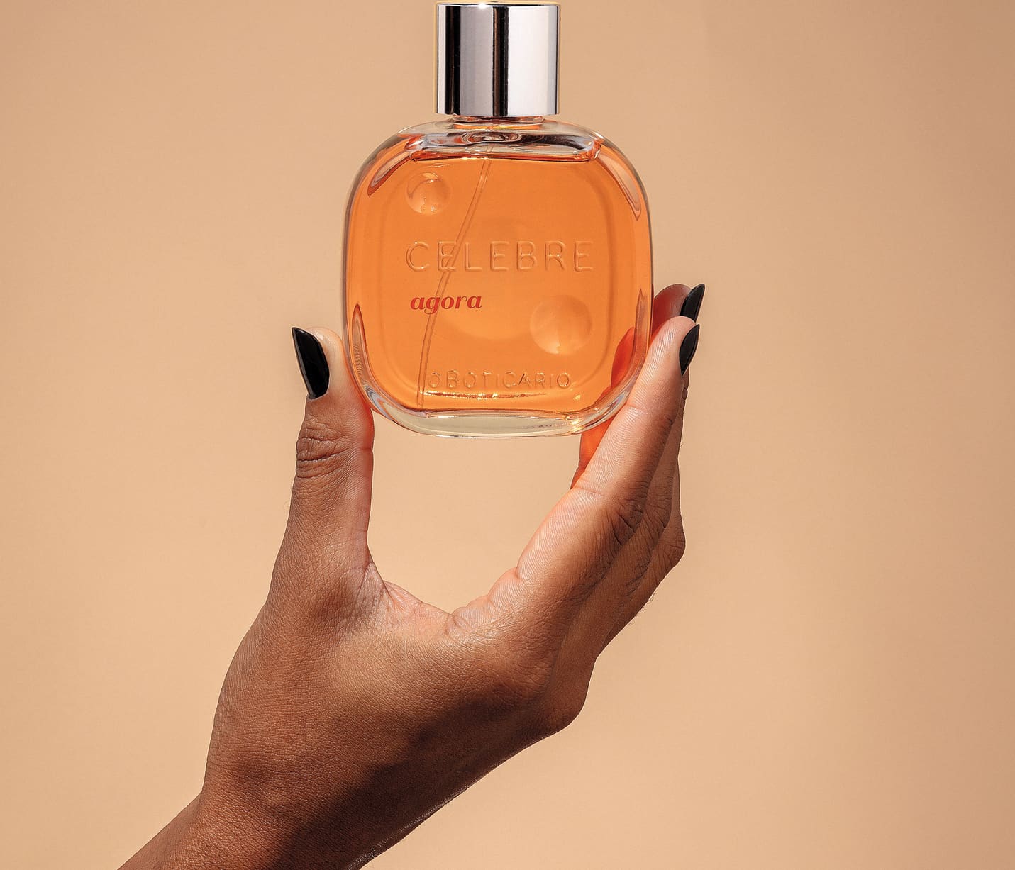 Uma mão feminina com unhas pintadas de preto segurando um frasco de vidro transparente com líquido laranja, tampa prateada e letras pretas e em relevo de desodorante colônia Celebre Agora Feminino em fundo em tons de laranja.