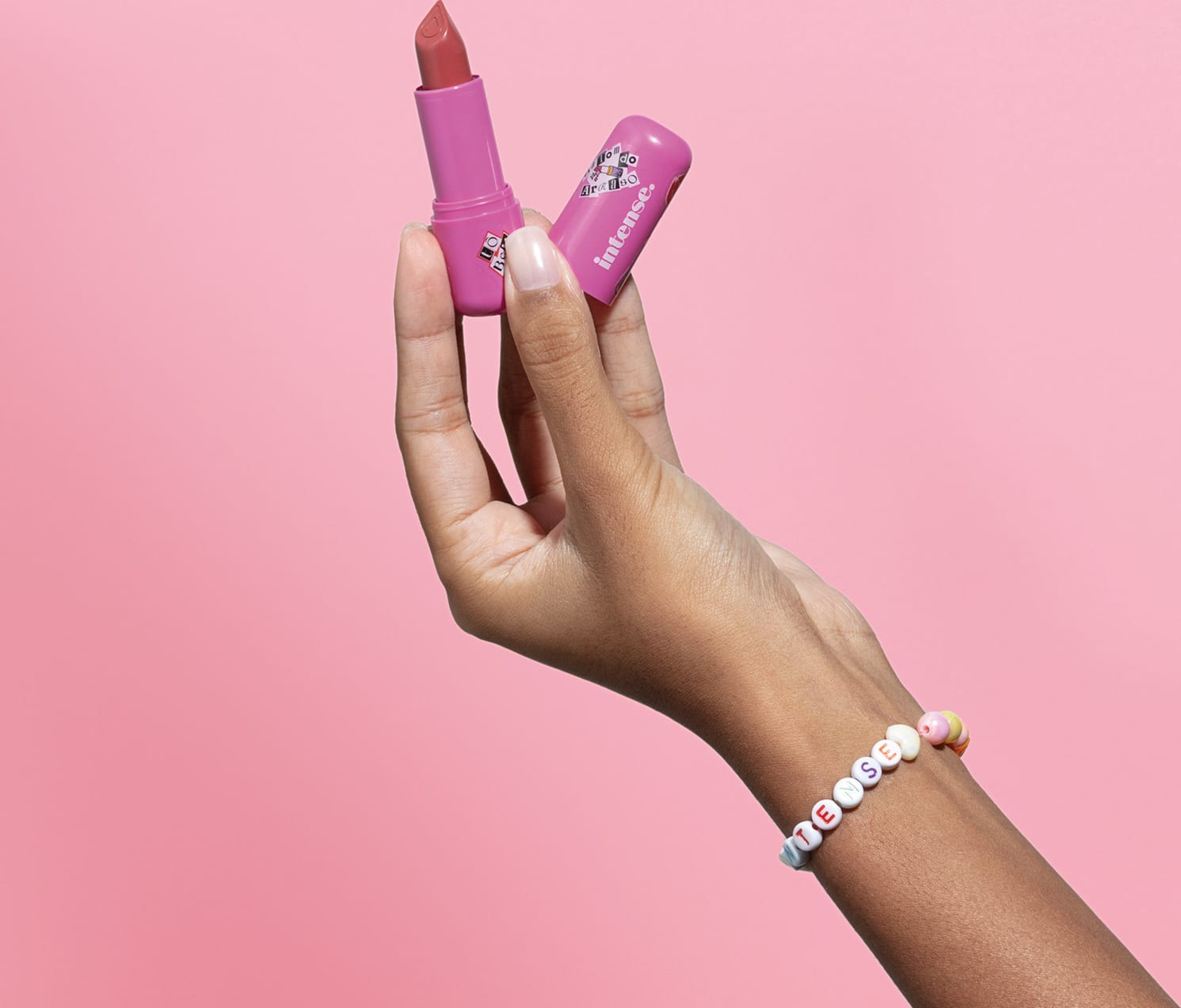 Uma mão feminina segura o batom do Arraso na cor pink rosé da marca Intense em embalagem na cor roxa e a outra mão segura a tampa, na mesma tonalidade, em fundo em tons de rosa.