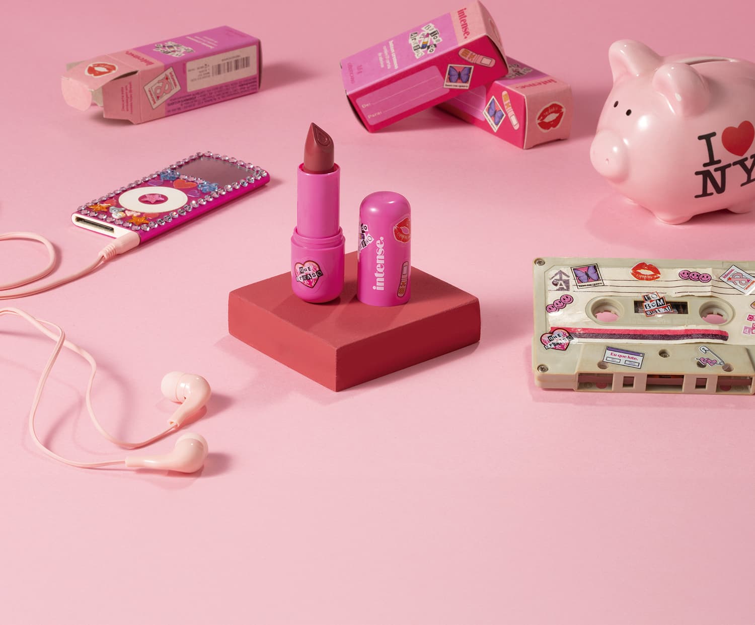 Batom do Arraso na cor pink rosé da marca Intense em embalagem na cor roxa está em cia de uma plataforma rosa com caixa do produto, mp3 player, cofre e fita cassete, tudo em tons de rosa.