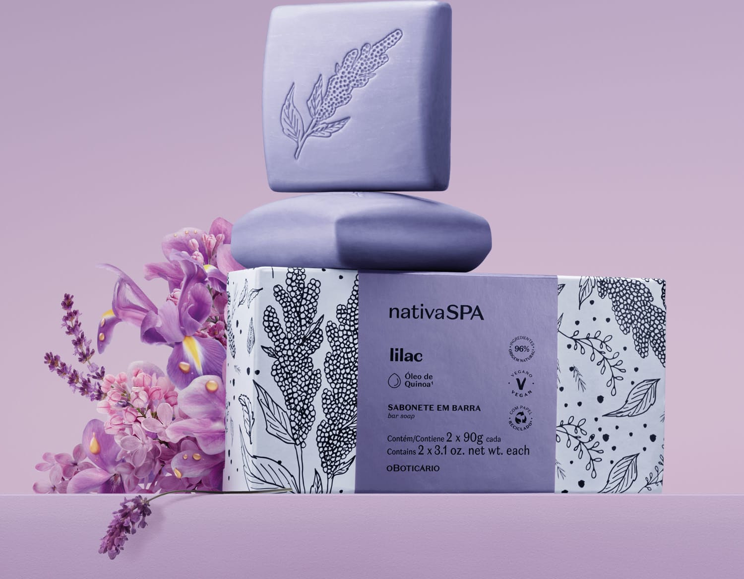Caixa branca com faixa lilás e ilustrações na cor preta ao lado de dois sabonetes em barra quadrados na cor lilás da marca Nativa SPA Lilac em um cenário com flores lilás ao redor e fundo na mesma tonalidade.