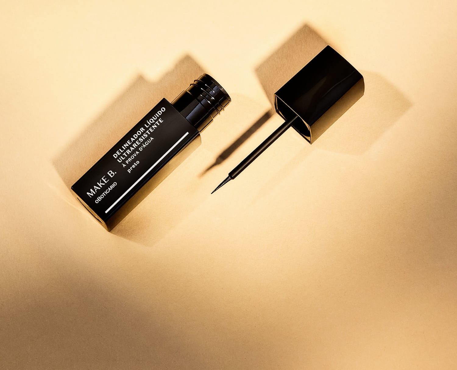 Tubo preto  com letras brancas de delineador líquido para olhos na cor preta da marca Make B., com tampa, haste e aplicador fino. A imagem tem um fundo em tons de rosa.