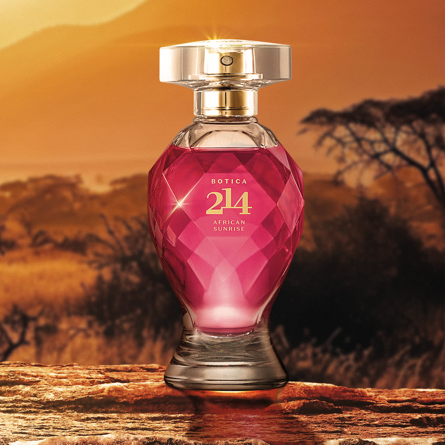 Vidro lapidado em formato de taça com líquido rosa vibrante, tampa transparente, pulverizador e letras douradas de Eau de Parfum Botica 214 African Sunrise Floral Frutal em cenário de paisagem natural com árvores ao fundo e céu alaranjado.
