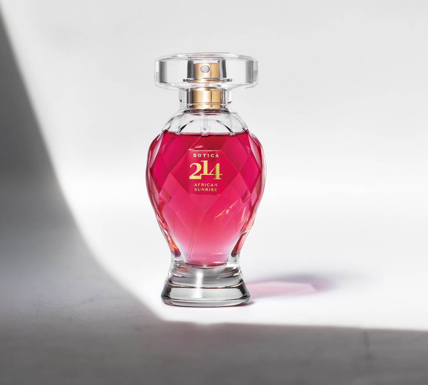 Vidro lapidado em formato de taça com líquido rosa vibrante, tampa transparente, pulverizador e letras douradas contendo 75ml de Eau de Parfum Botica 214 African Sunrise Floral Frutal em fundo claro com sombras.