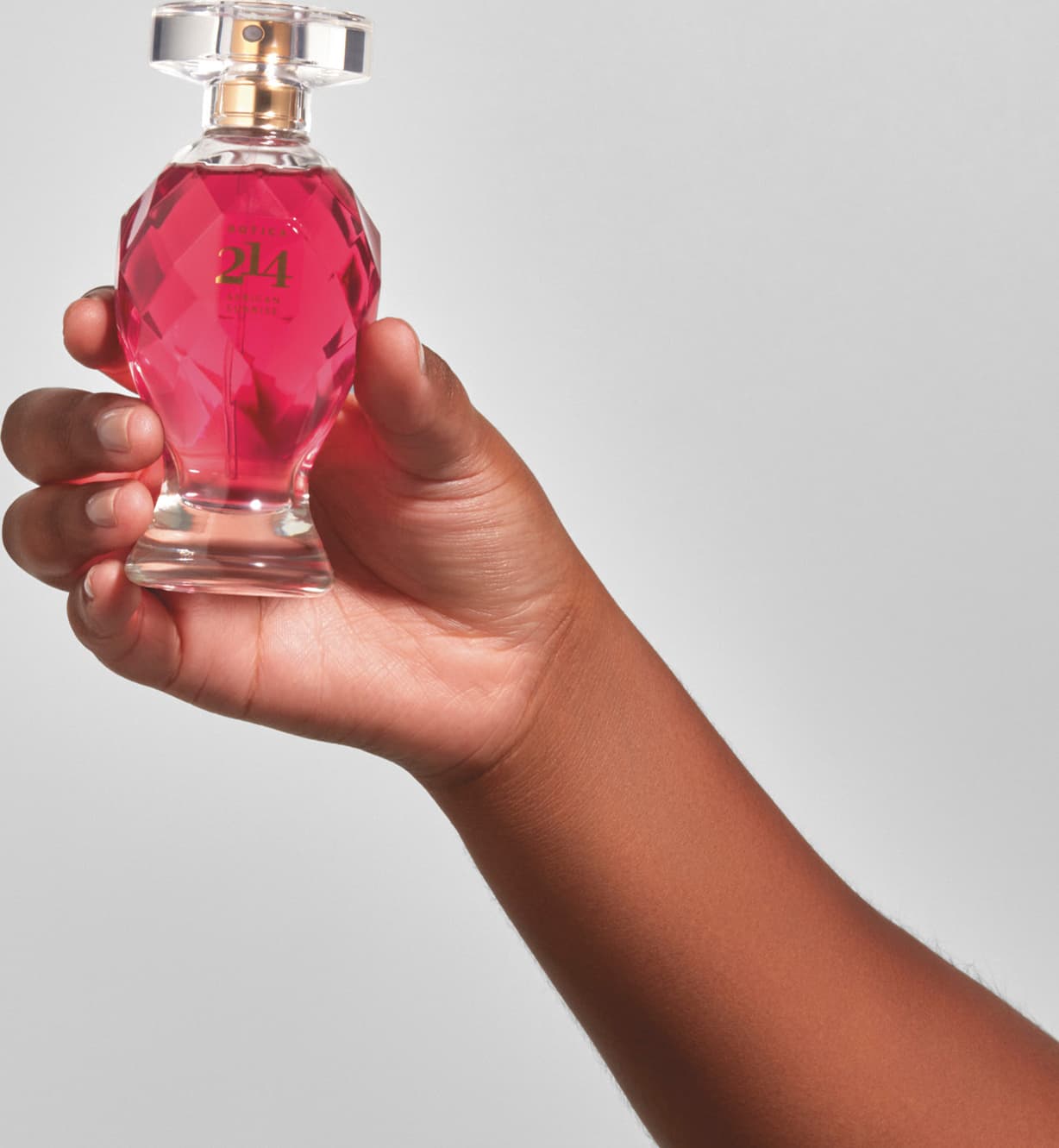 Mão feminina segurando um vidro lapidado em formato de taça com líquido rosa vibrante, tampa transparente, pulverizador e letras douradas contendo 75ml de Eau de Parfum Botica 214 African Sunrise Floral Frutal em fundo azul.