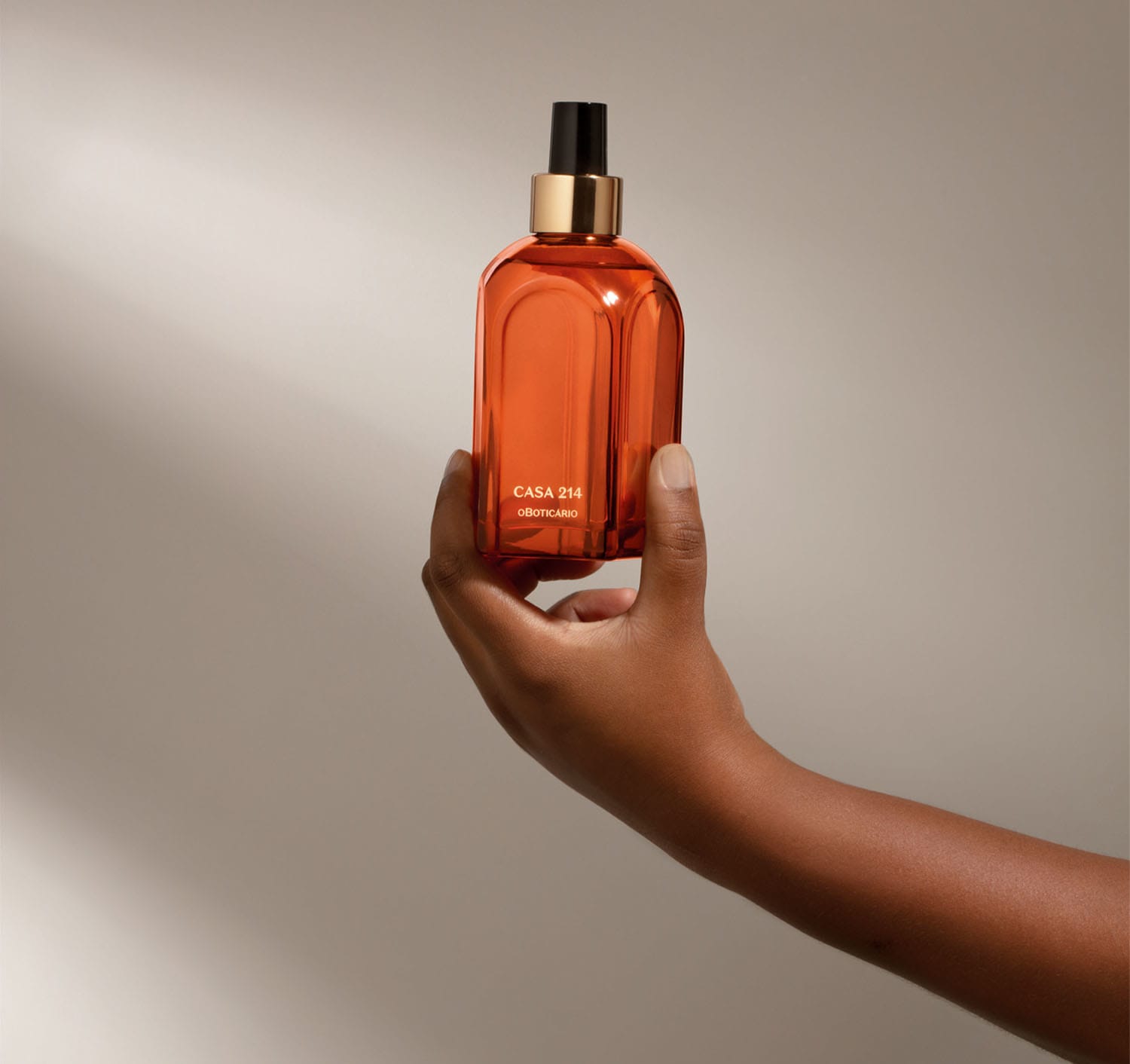 Mão segurando um vidro laranja de tampa spray preta com detalhe e letras douradas contendo aromatizador de ambientes Casa 214 Vanilla Sublime em fundo cinza com sombras.