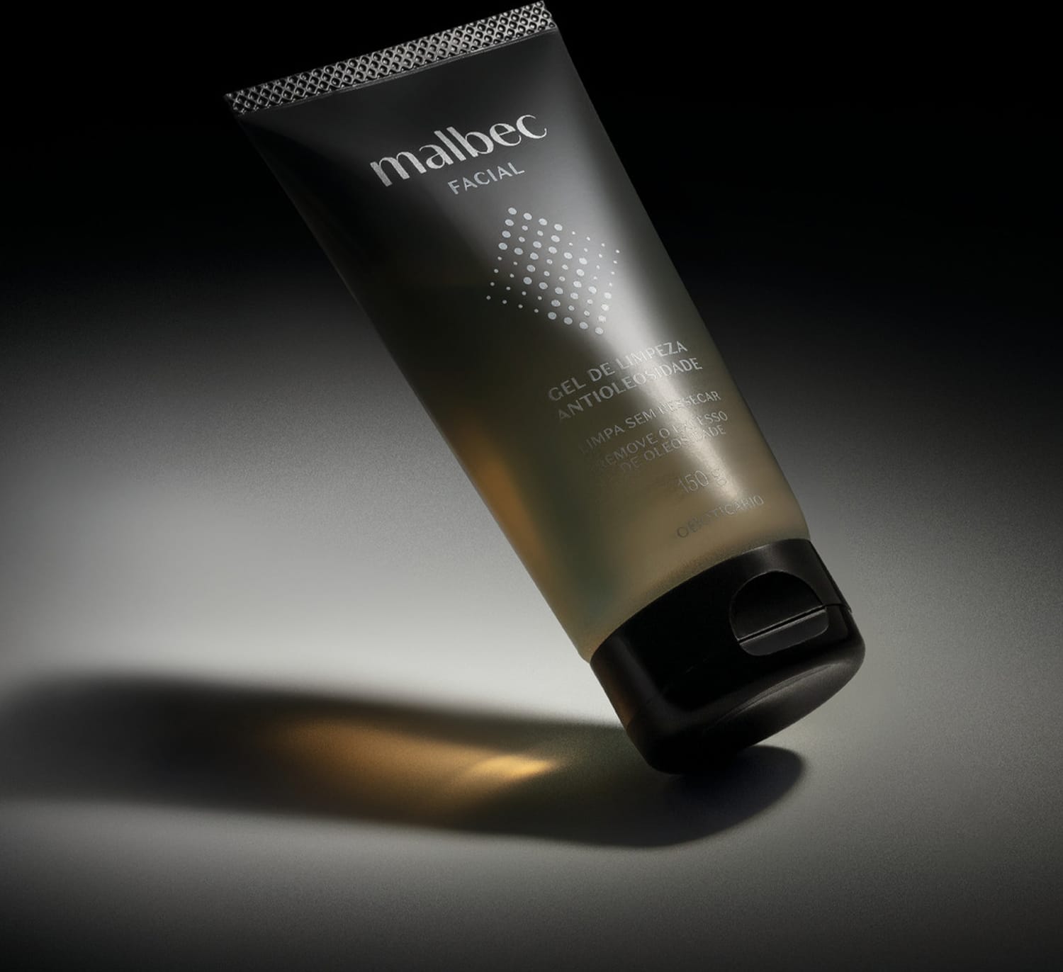 Frasco com degradê de preto e branco com tampa preta e rótulo com letras e detalhes brancos contendo 150g de gel de limpeza antioleosidade Malbec Facial em fundo escuro com pouca iluminação.