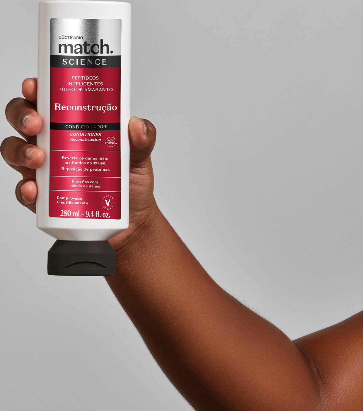 Mão segurando um vidro branco com tampa preta e rótulo vermelho, preto e prateado de condicionador Match Science Reconstrução em fundo cinza.