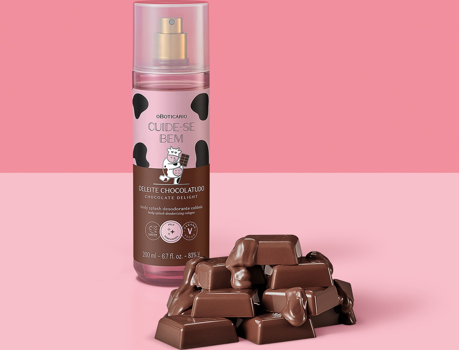 Frasco com rotulo rosa e marrom com vaca ilustrada de body splash Cuide-se Bem Deleite Chocolatudo com pedaços de chocolate ao leite espalhados ao lado do frasco, representando o aroma do produto em fundo rosa.