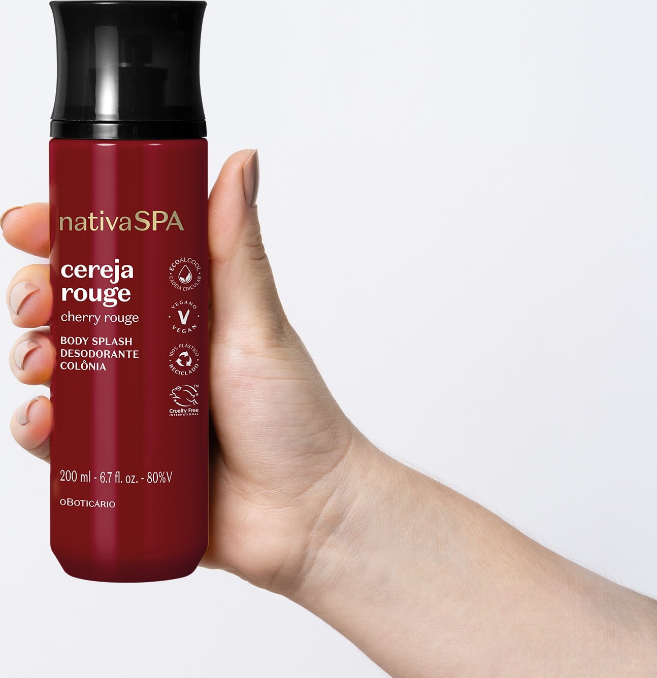 Mão feminina segurando um frasco vermelho escuro com tampa preta de body splash Nativa SPA Cereja Rouge em fundo azul claro.