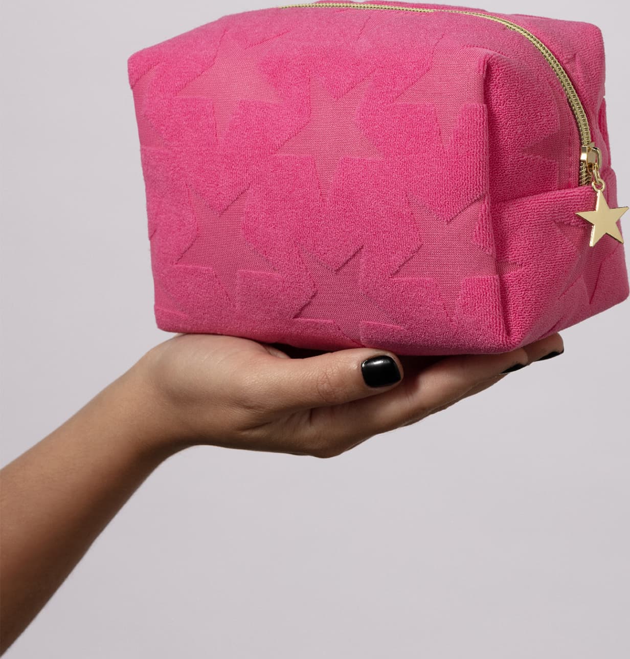 Mão segurando um nécessaire rosa macio com estampa de estrelas e fecho em zíper com pingente também de estrelas em fundo cinza claro.