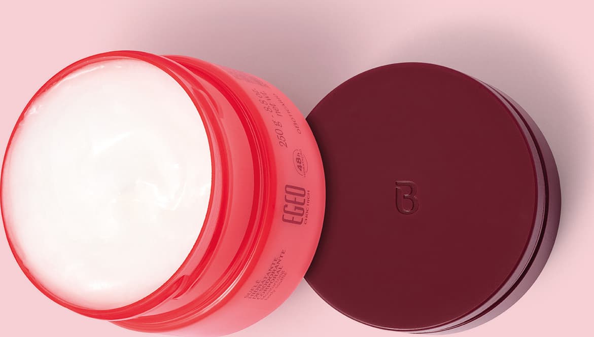 Pote aberto de Suflê Hidratante Desodorante Corporal da linha Egeo Choc High. A tampa vinho está sobre uma superfície rosa ao lado da embalagem, vistos de cima, o que permite ver o creme corporal branco dentro do pote rosa.