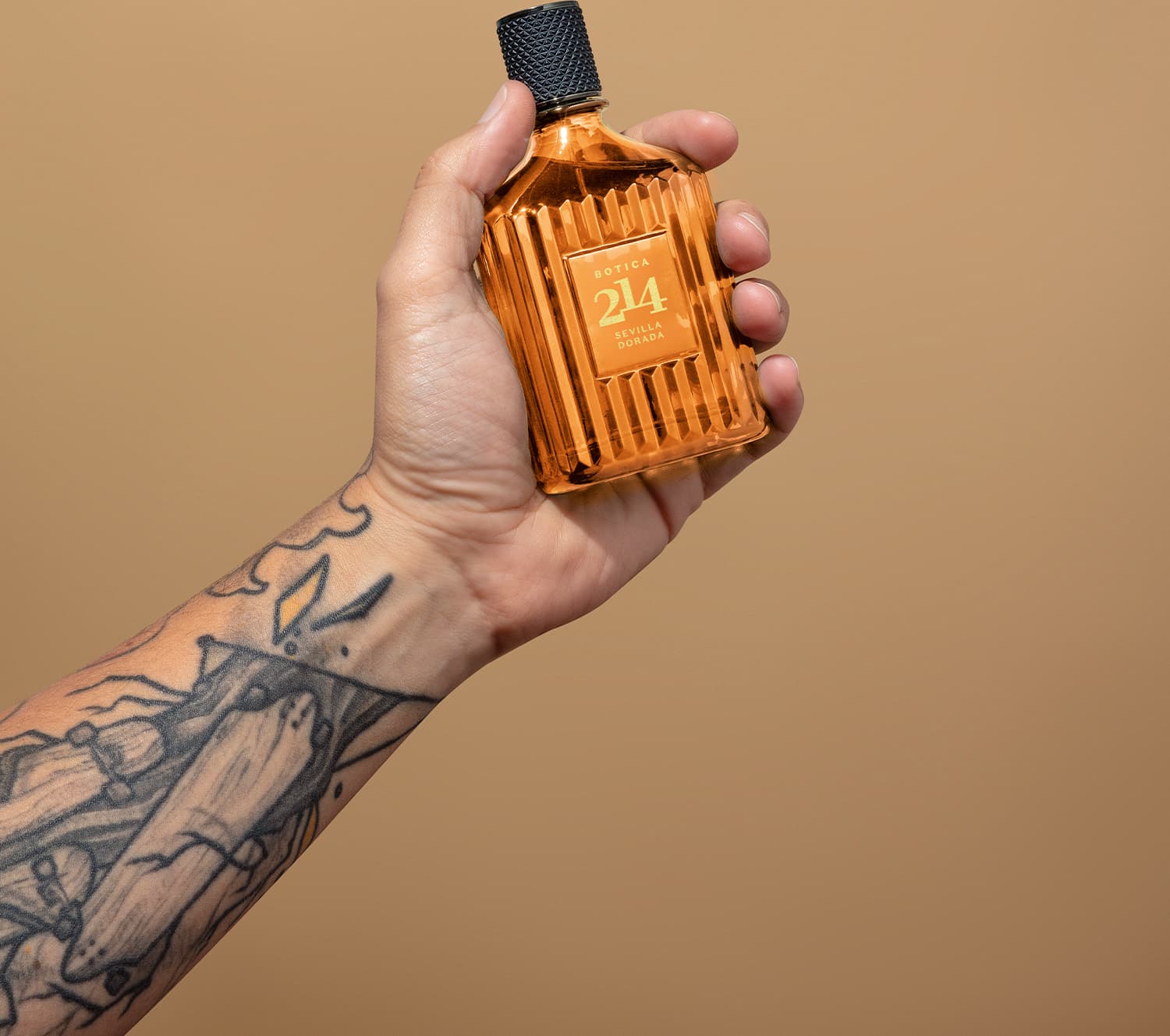 Frasco de Eau de Parfum Fougére Aromático Botica 214 Sevilla Dorada em mão com braço tatuado, sobre fundo bege, em ambiente minimalista e monocromático.