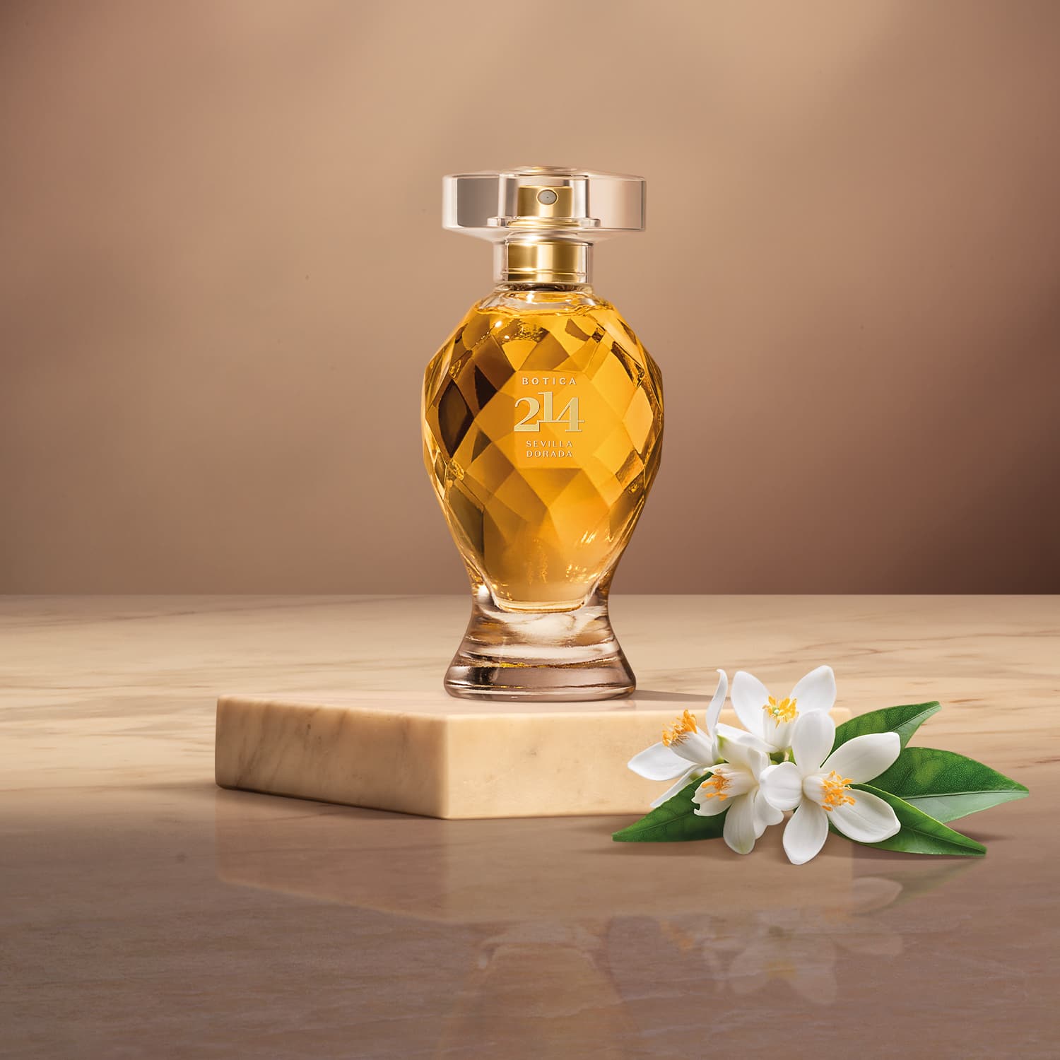 Frasco de Eau de Parfum Floral Frutal Botica 214 Sevilla Dorada com design facetado em âmbar, sobre superfície de madeira clara, com flor branca e folhas verdes ao lado.