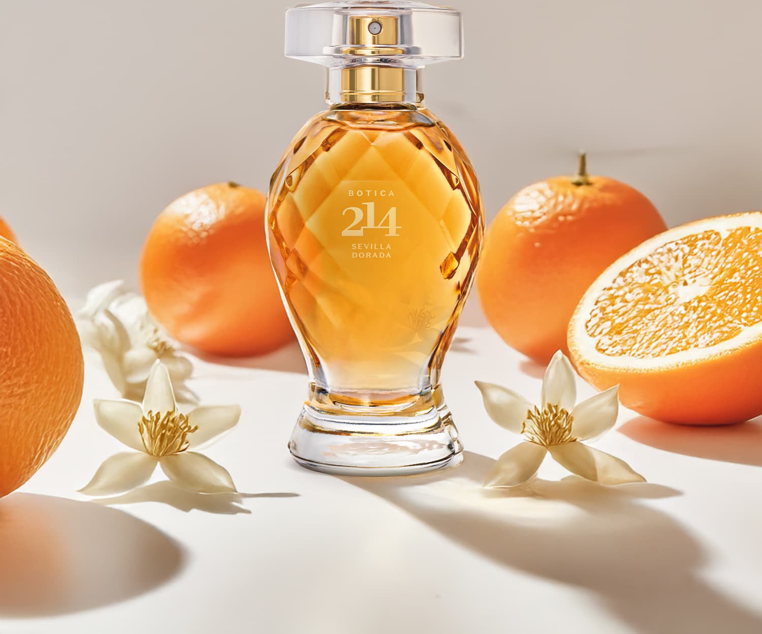 Frasco de Eau de Parfum Floral Frutal Botica 214 Sevilla Dorada em meio a laranjas frescas e flores brancas sobre superfície clara, em cenário iluminado e com cores quentes predominantes.