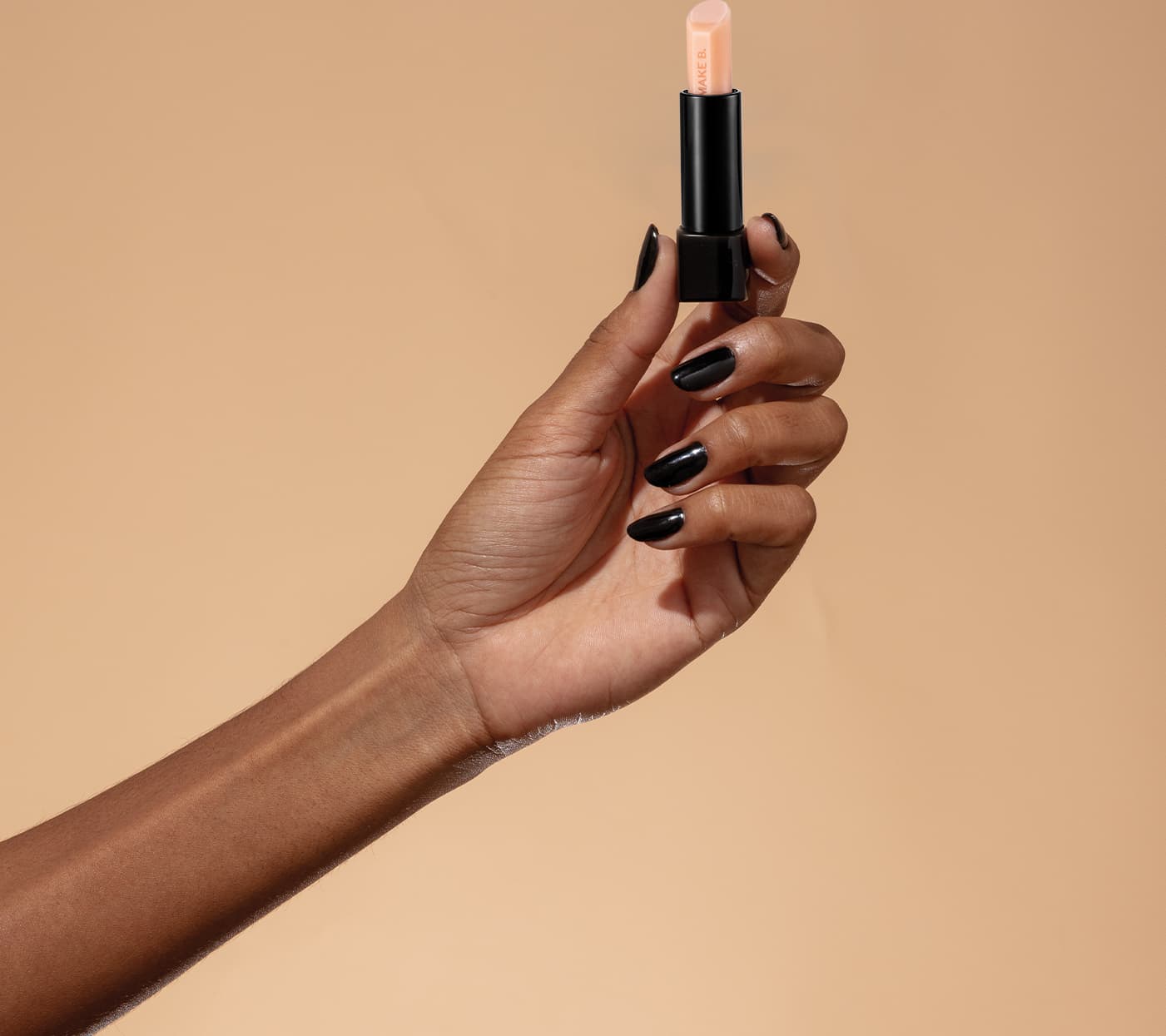 Mão feminina com unhas pintadas de esmalte escuro segurando um balm labial noturno da marca Make B. Lip Chronology Night Plump sobre fundo bege.