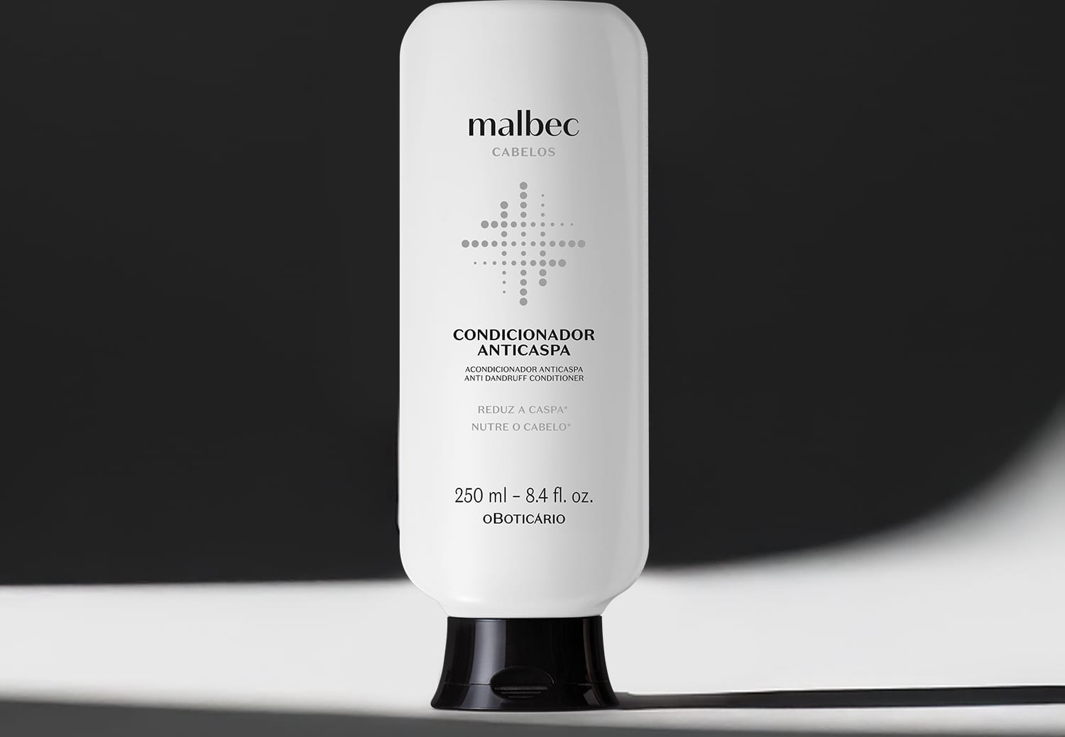 Imagem de frasco branco com tampa preta e rótulo com letras pretas da marca Malbec para cabelos, sobre superfície branca iluminada com luz cênica projetando uma sombra a direita do frasco sobre fundo preto.