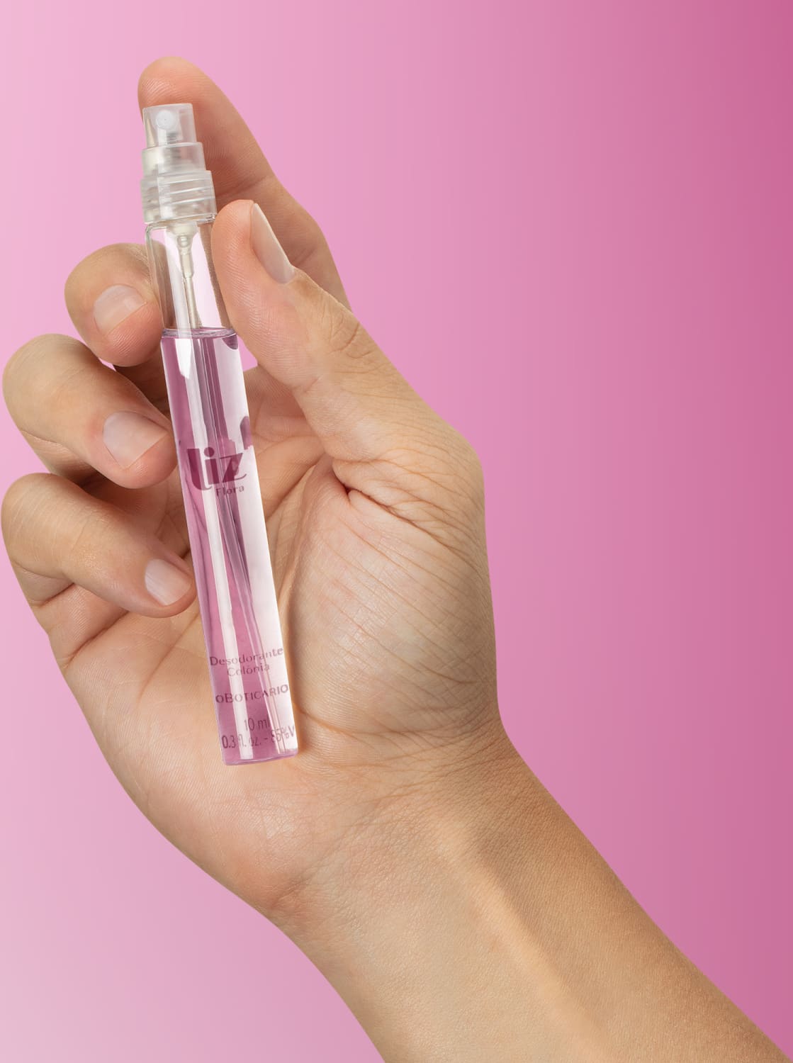 Mão feminina segurando um frasco cilíndrico transparente com líquido claro, tampa preta e letras na cor vinho contendo 10ml de desodorante colônia Liz Flora em fundo em tons de roxo.