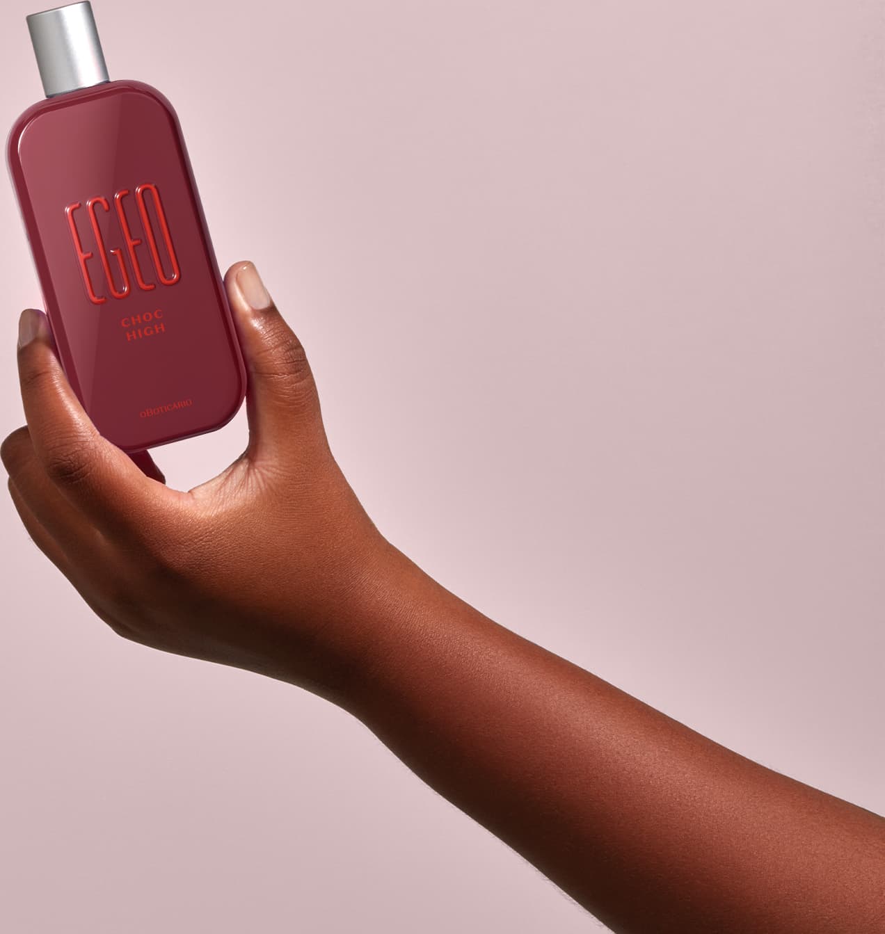 Frasco vermelho de desodorante colônia Egeo Choc High sendo segurado por mão feminina de pele negra contra fundo rosa claro.