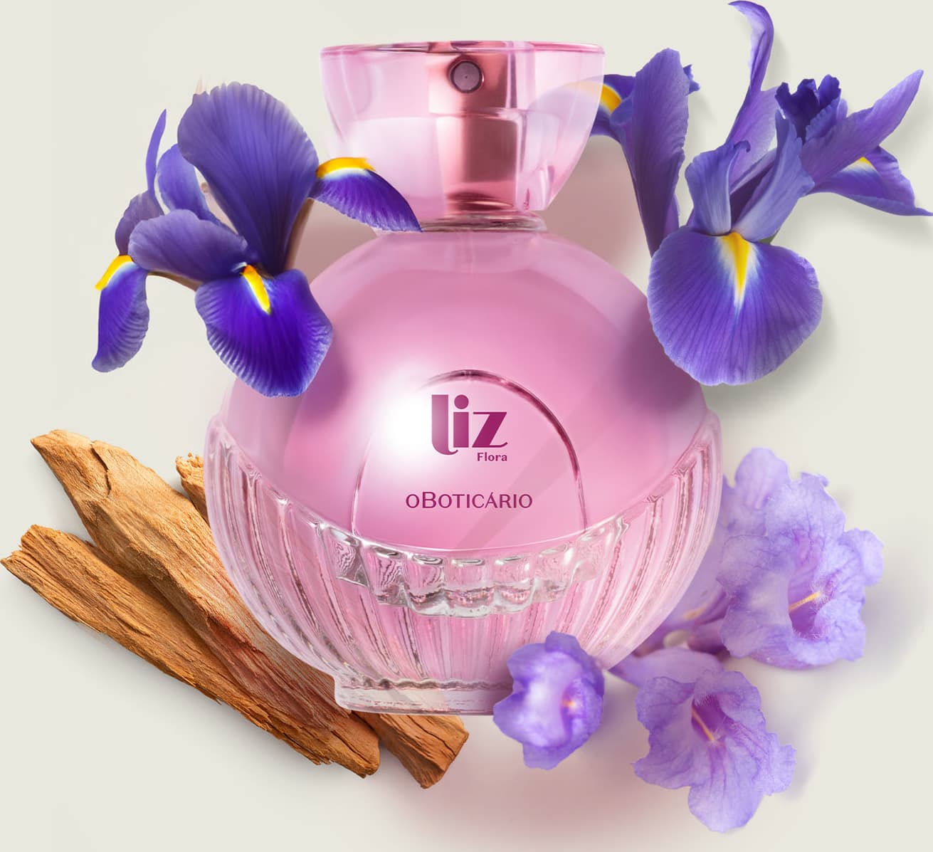 Vidro arredondado com líquido rosa, letras vinho, borrifador dourado e tampa transparente contendo desodorante colônia Liz Flora rodeado por flores de íris violetas e pedaços de madeira em superfície clara.