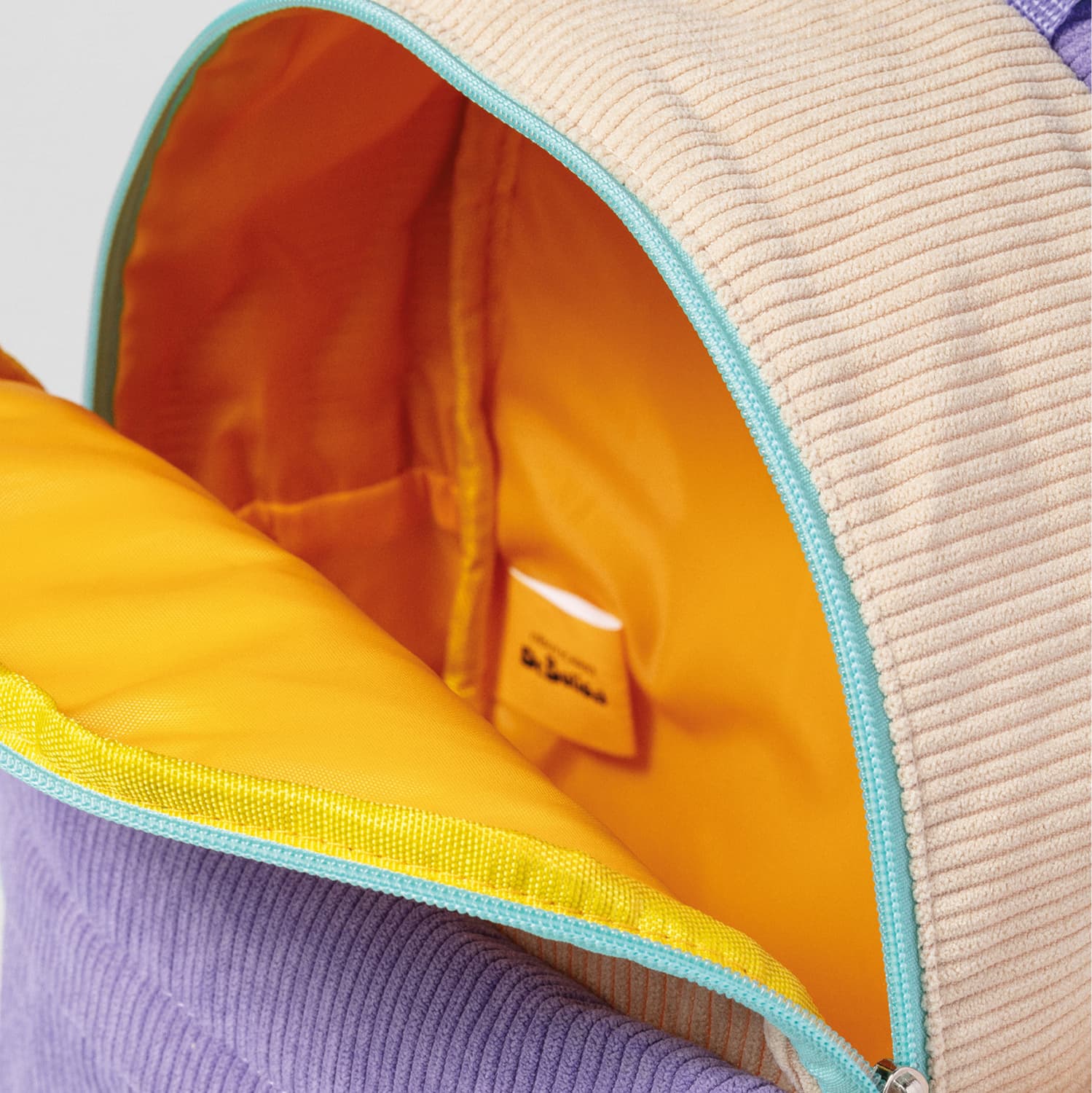 Close-up de mochila infantil de tecido macio em tons de bege e roxo, com zíper azul-turquesa por fora. A mochila está aberta e seu interior é amarelo intenso., 