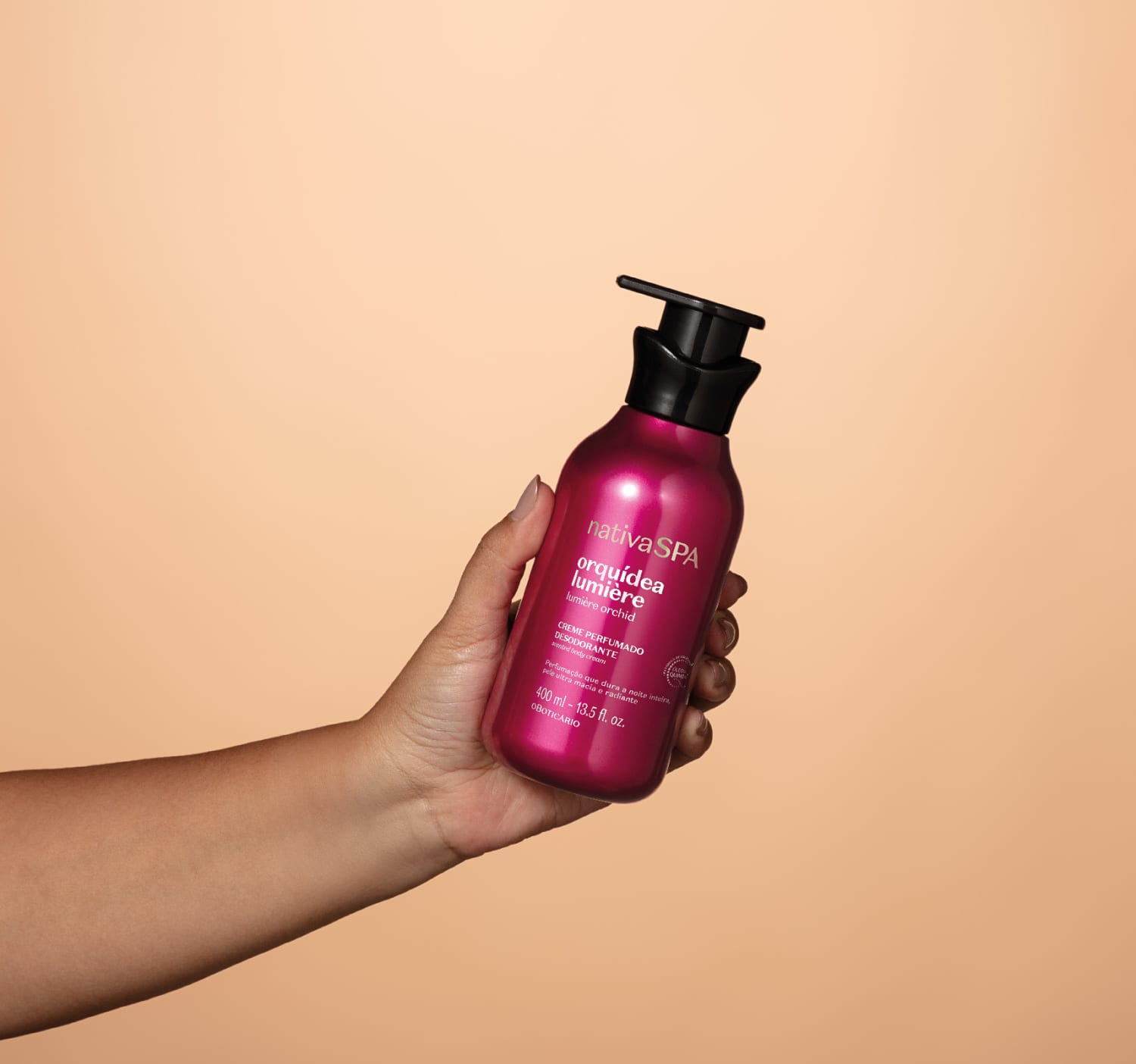 Embalagem pump rosa brilhante com tampa preta de item de corpo e banho da marca Nativa SPA Orquídea Lumiere sendo segurada por mão feminina em fundo laranja claro.