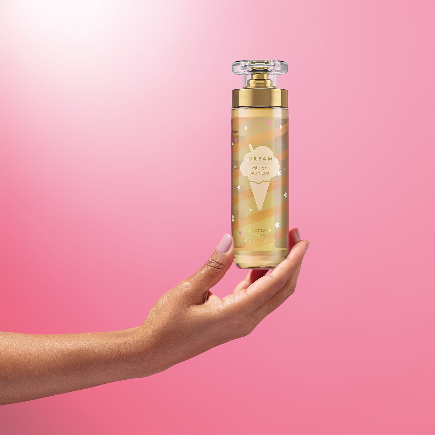 Imagem de uma mão segurando um frasco de body splash Dream Céu de Baunilha. A embalagem conta com tampa transparente, válvula spray dourada e rotulo em tons de dourado. O cenário traz um fundo em tons de lilás e branco.