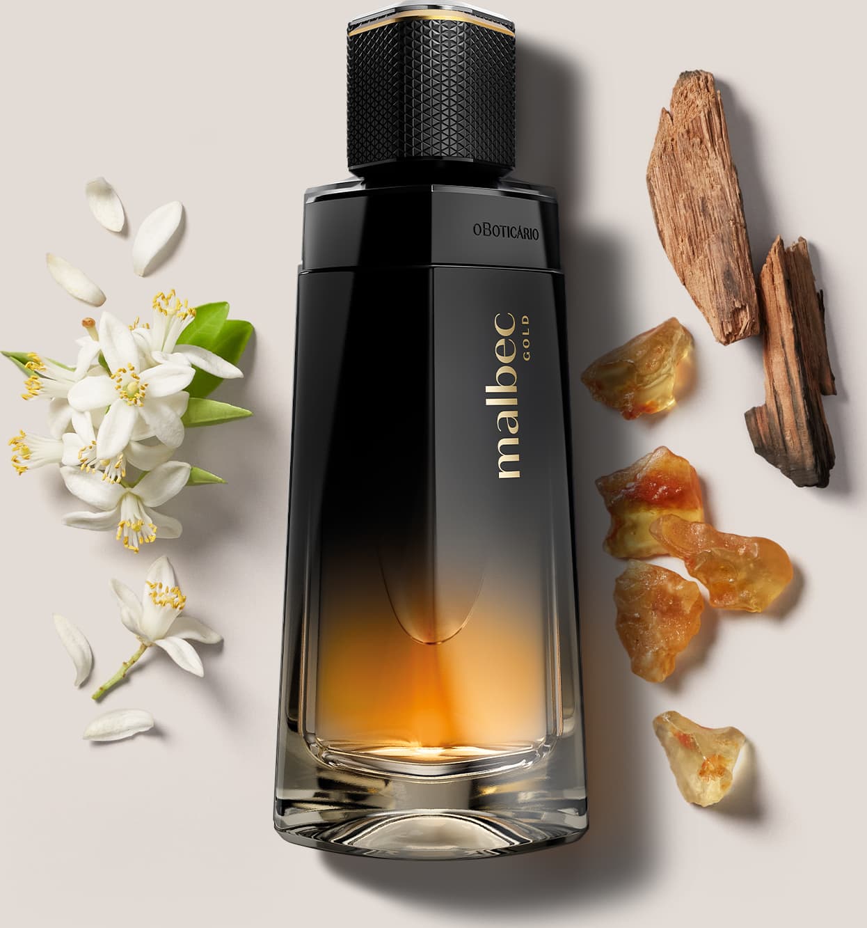 Frasco de desodorante colônia Malbec Gold nas cores degradê de preto com dourado cercado por elementos naturais, como flores brancas e pepitas de âmbar e madeiras, sugerindo ingredientes naturais que compõem a fragrância.