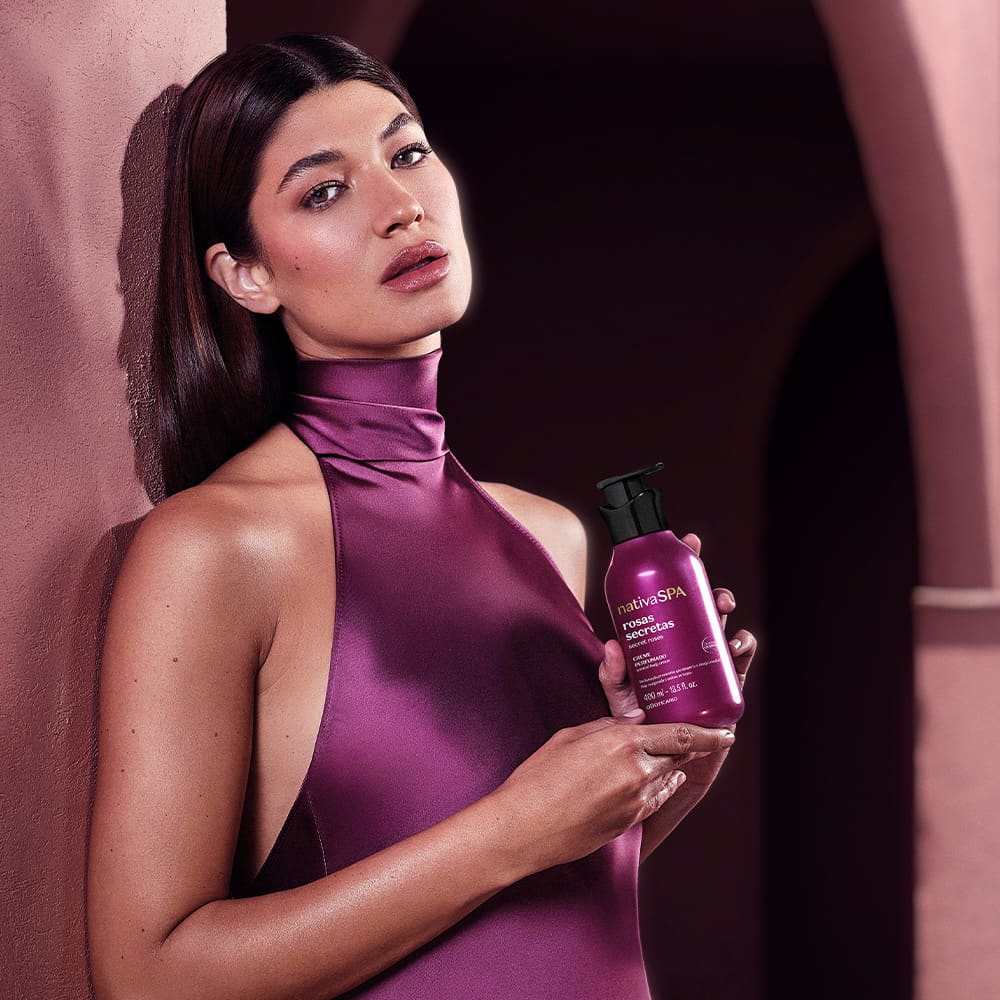 Frasco de creme corporal Nativa SPA, na cor roxa, com rótulo destacando o nome "Rosas Secretas". O produto é segurado por uma mulher com pele branca, cabelos escuros e vestido roxo, em um cenário com parede lilás.