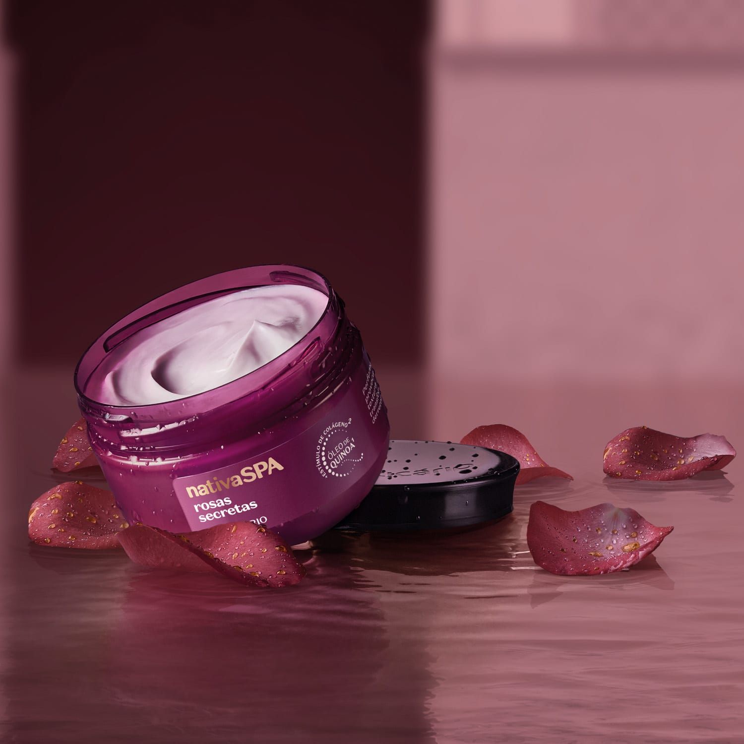 Ambiente de spa com um pote de óleo em creme Nativa SPA Rosas Secretas. Ele está sobre pétalas de rosa espalhadas sobre uma superfície úmida, em um cenário monocromático de tons de roxo.