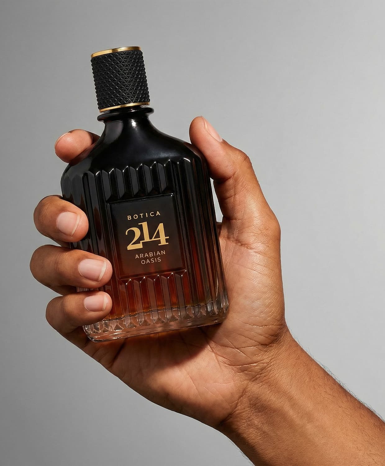 Frasco de item de perfumaria da marca Botica 214 Arabian Oasis sendo segurado por mão masculina sobre fundo cinza, com textura canelada e logo gravado em dourado no vidro da embalagem.